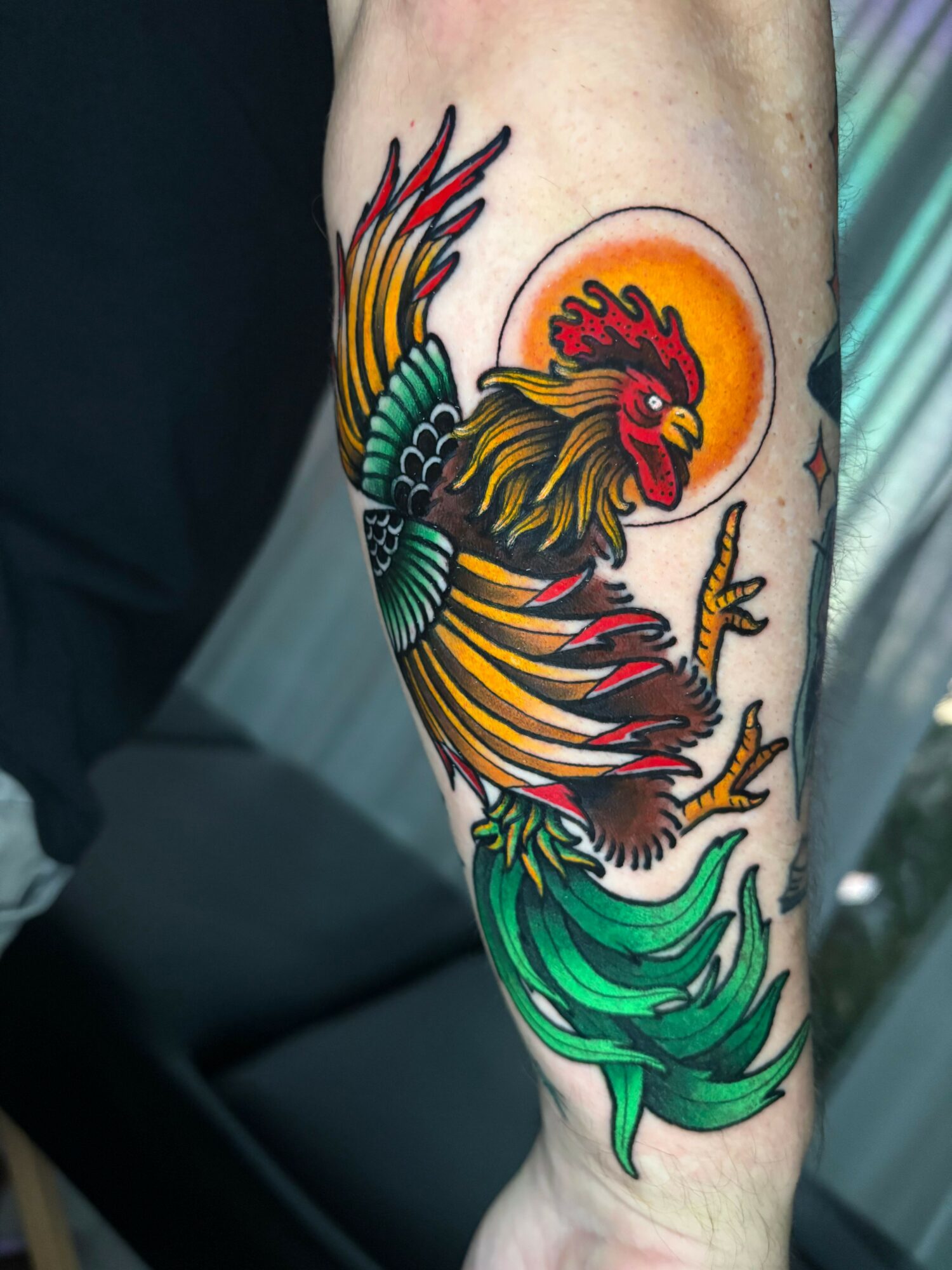 Colorful rooster tattoo with a sun background on person's forearm, featuring detailed feathers and green tail feathers.