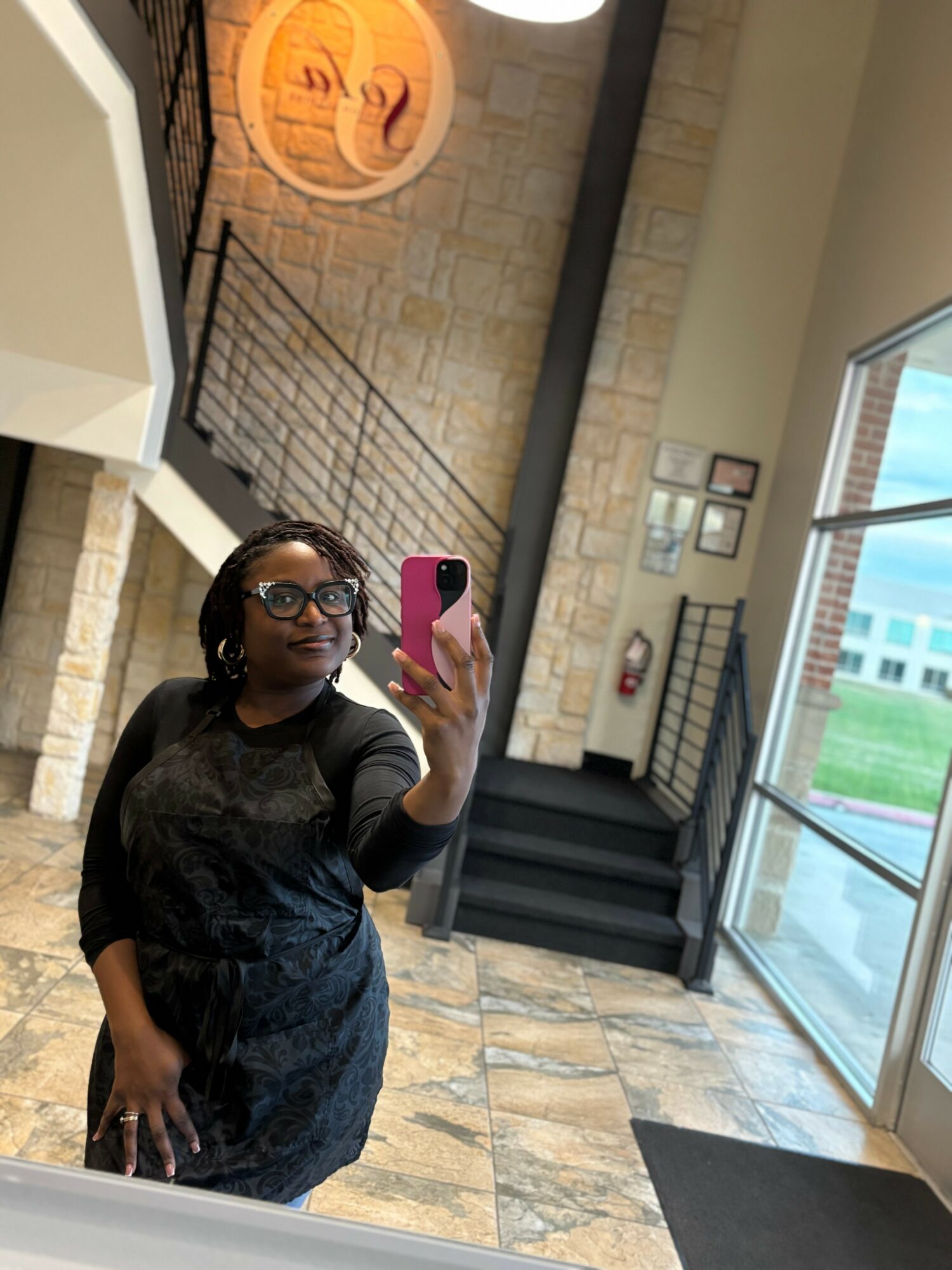 Woman taking a mirror selfie indoors, wearing glasses and a dark dress, with stairs and large windows in background.