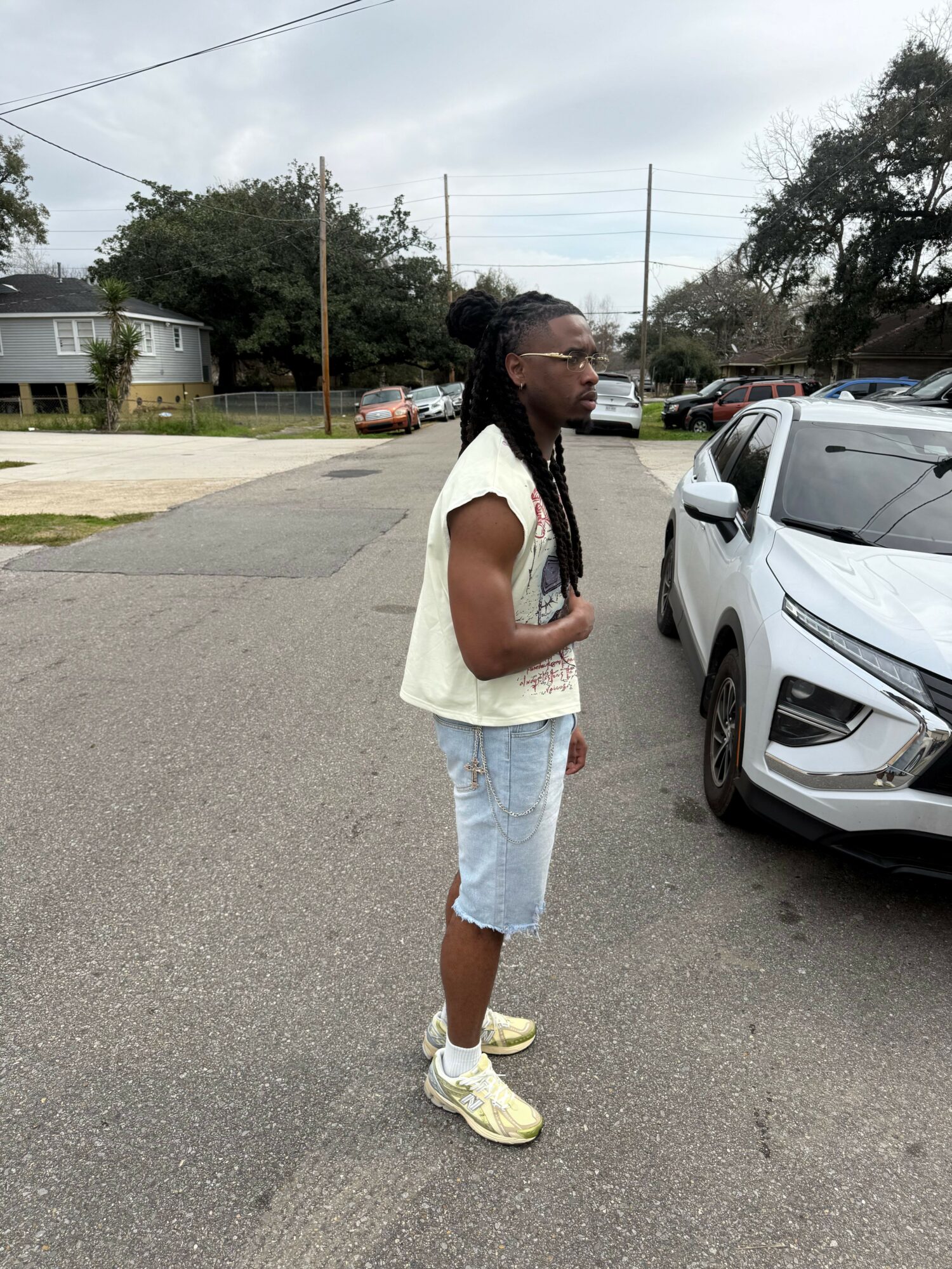 Person with long braided hair, glasses, white sleeveless top, and light blue shorts standing beside a white car.