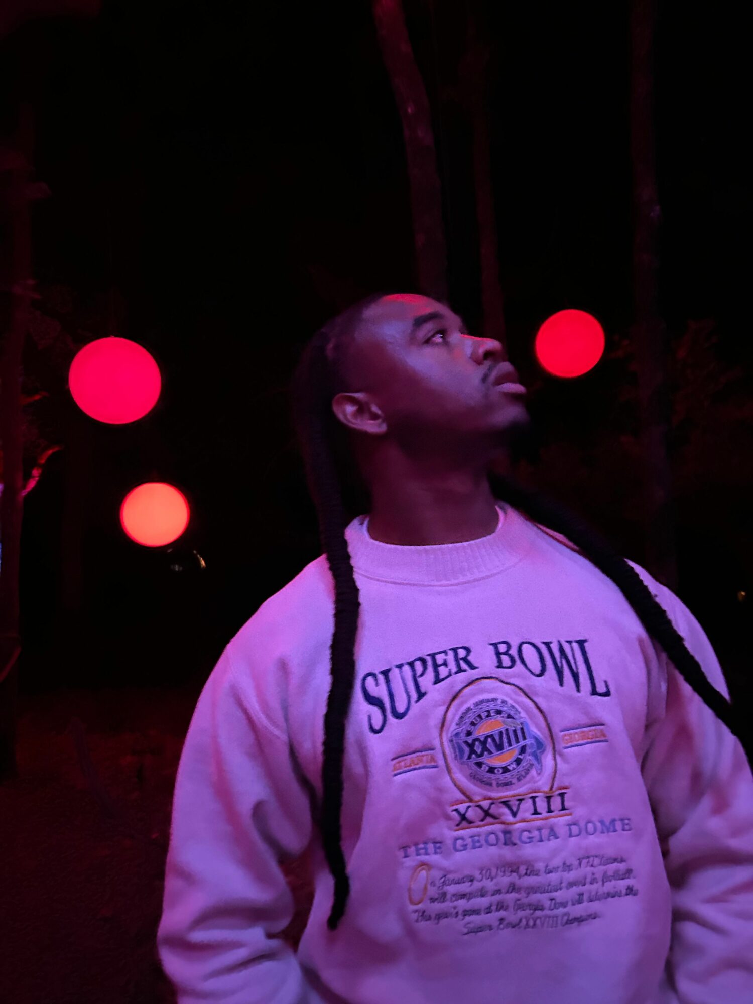 Person with long braids wearing a Super Bowl sweatshirt, looking upward in a dark setting with red lights.
