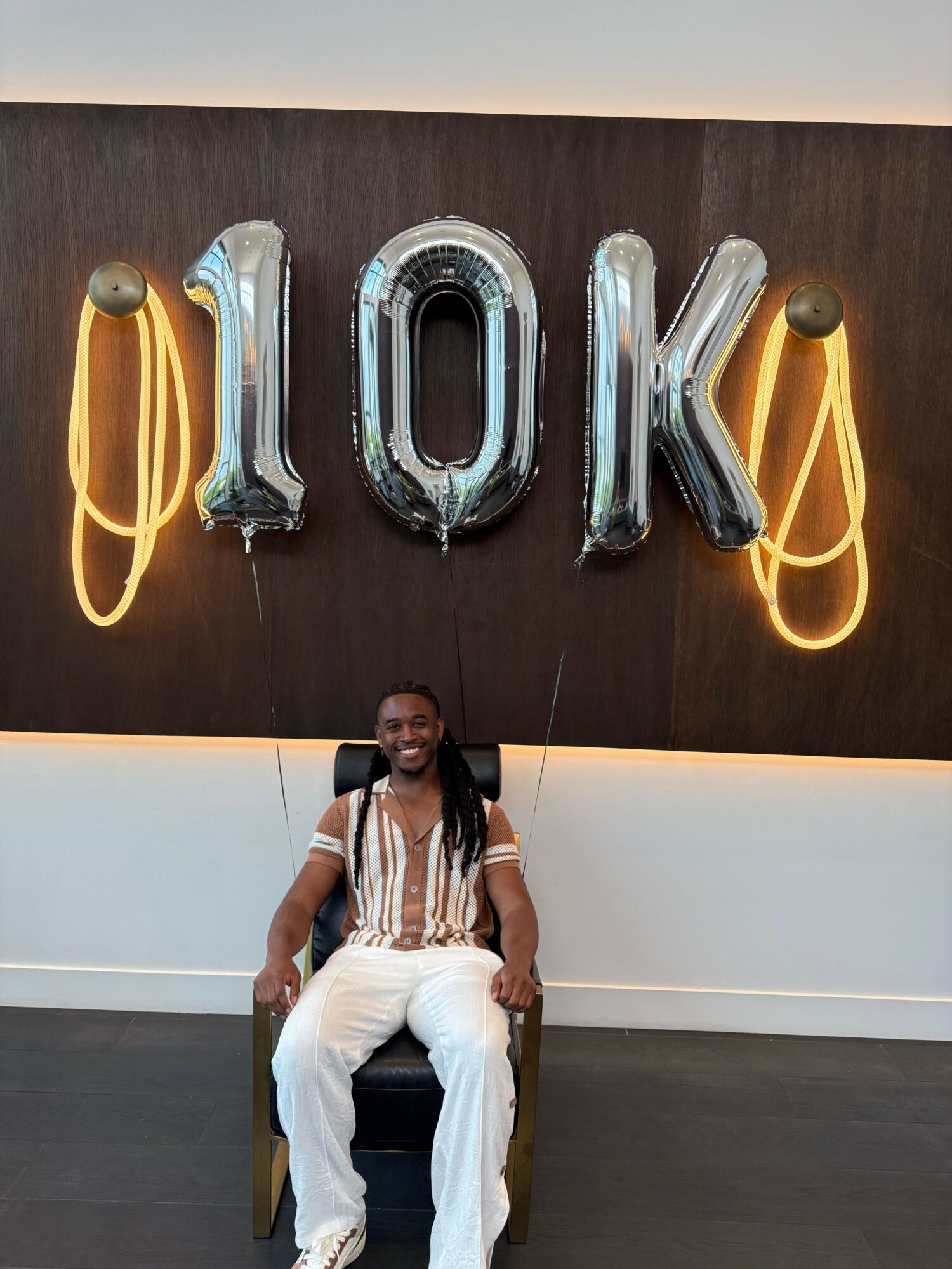 Person sitting on a chair in front of large silver balloons spelling '10K' with yellow ribbons, against a dark wall.
