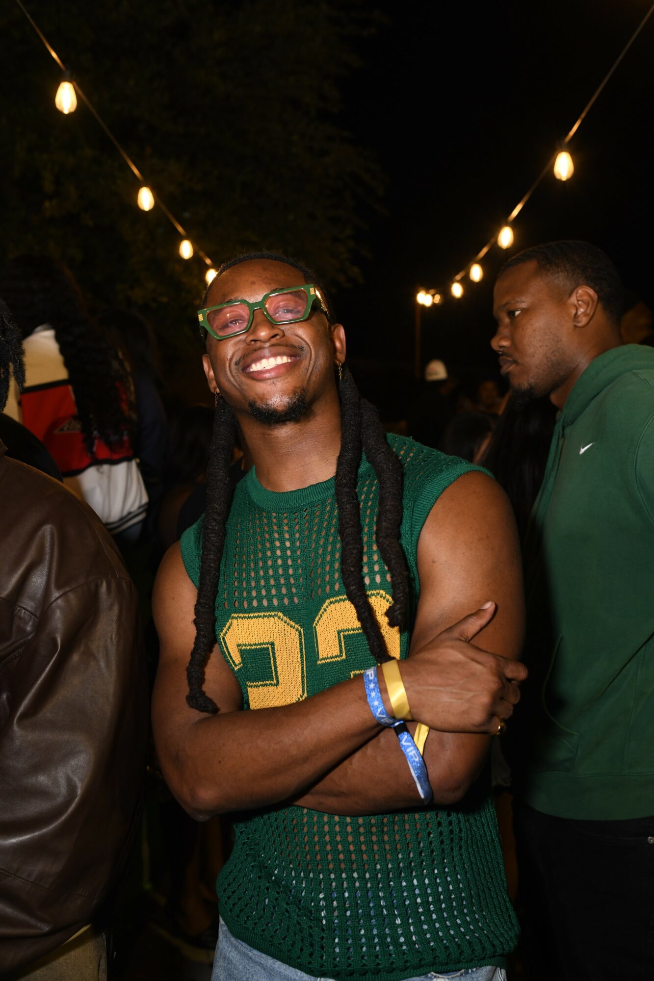 Smiling man with glasses and long braids at outdoor night event, wearing green sports jersey, surrounded by people and string lights.
