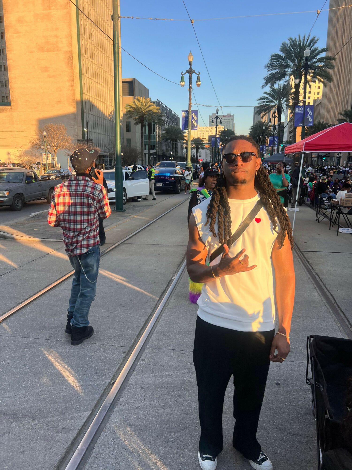 Young woman with long hair, sunglasses, white t-shirt, black pants, standing near tram tracks on city street.
