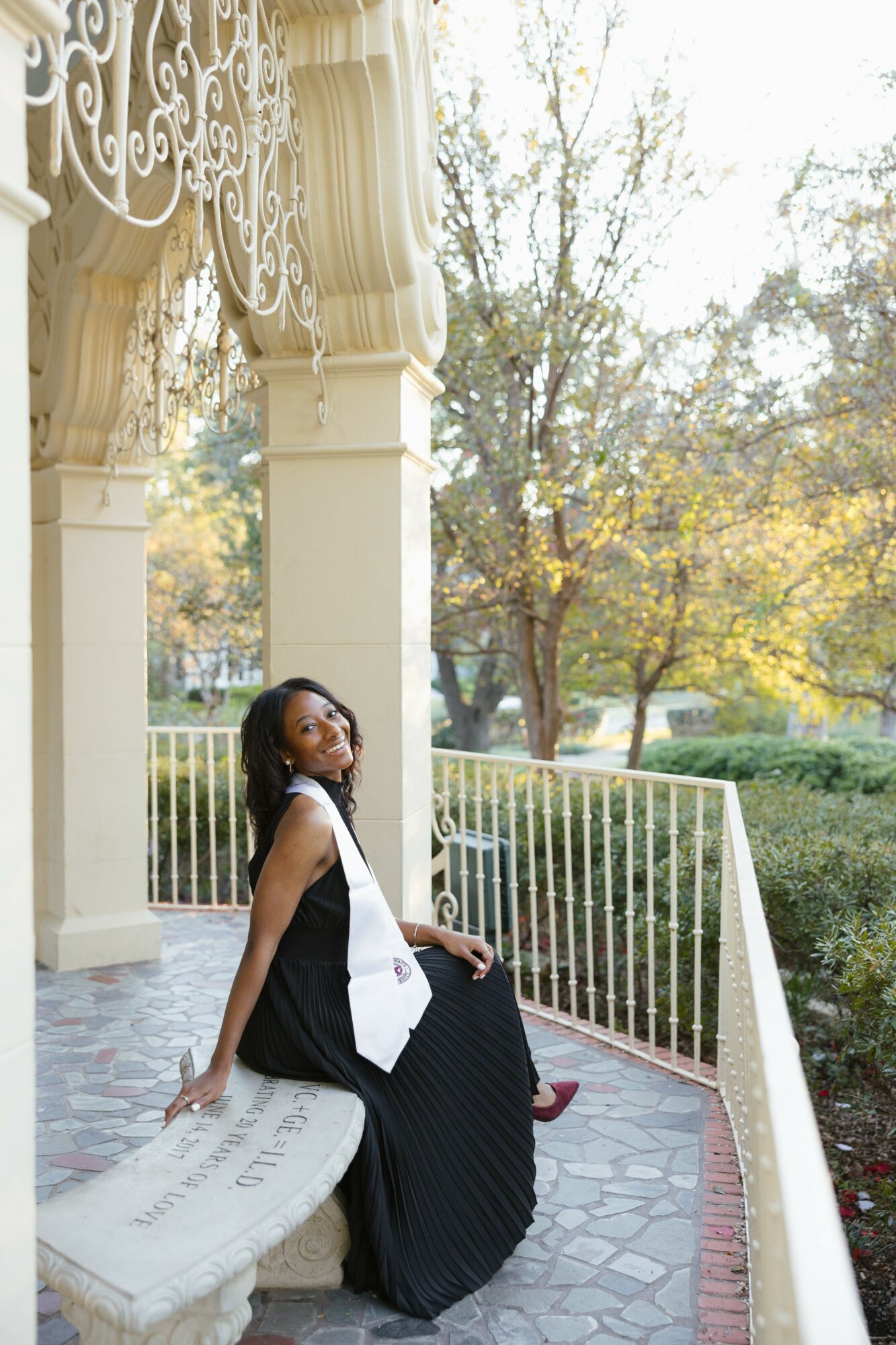 Woman in black dress sitting on a stone bench outdoors with trees and a white railing behind her.