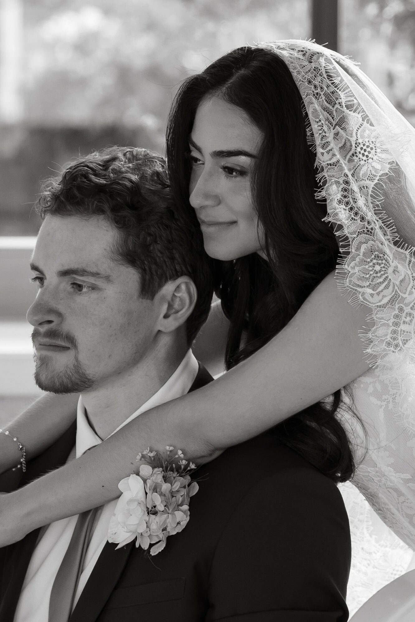A woman with dark hair and a lace veil leans on a man with short hair and a beard, both in formal attire.