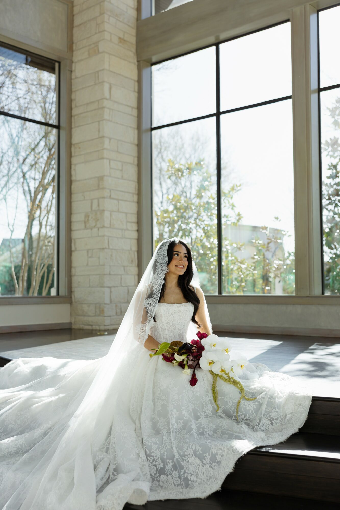Bride in wedding dress and veil sitting on floor near large windows with trees outside.