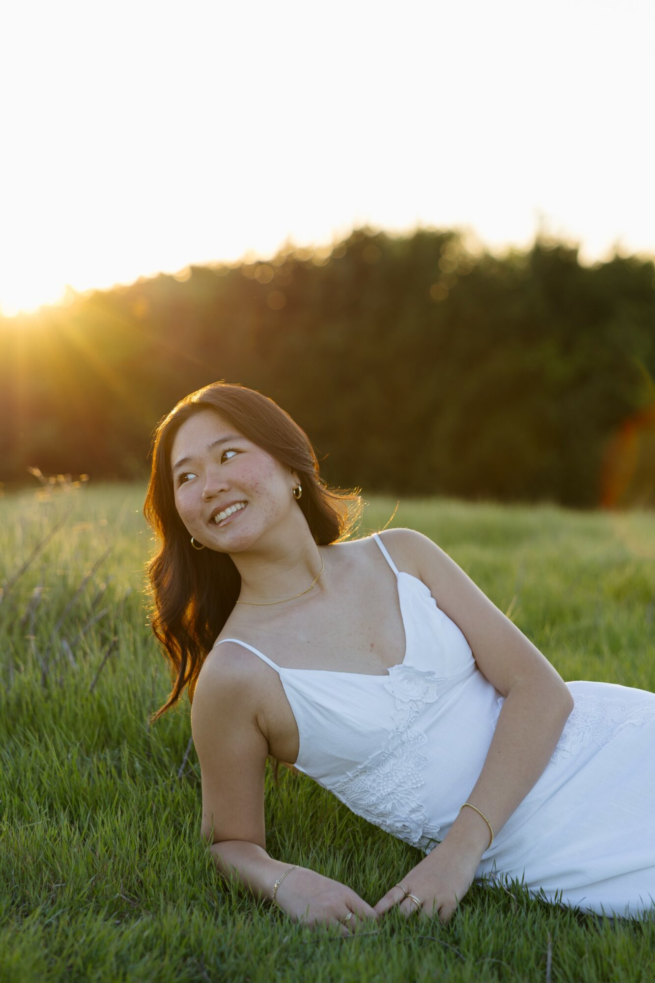 Woman lying on grass in a white dress, smiling, with trees and sunset in background.