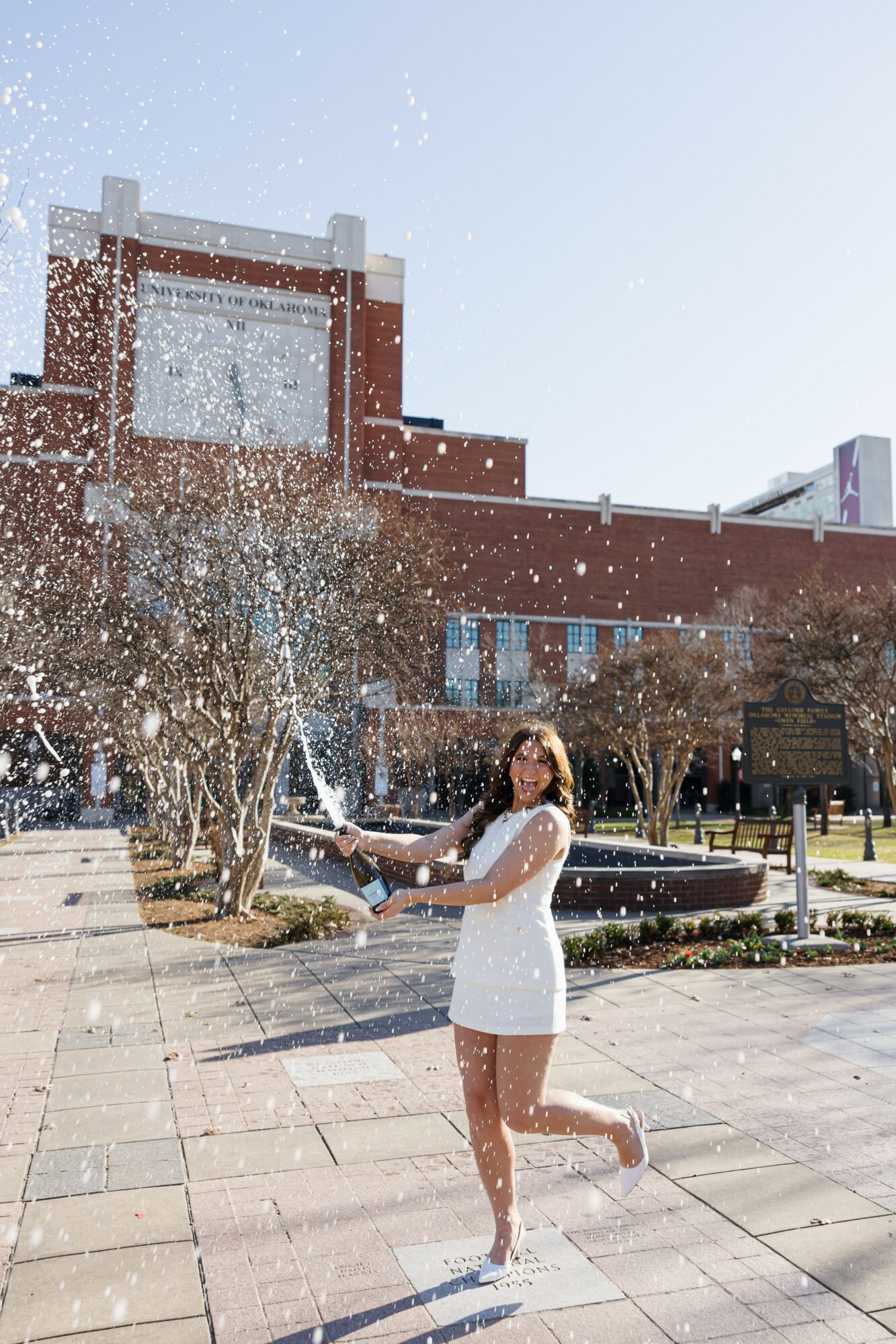 Woman in white dress standing outdoors with trees and a brick building in the background.