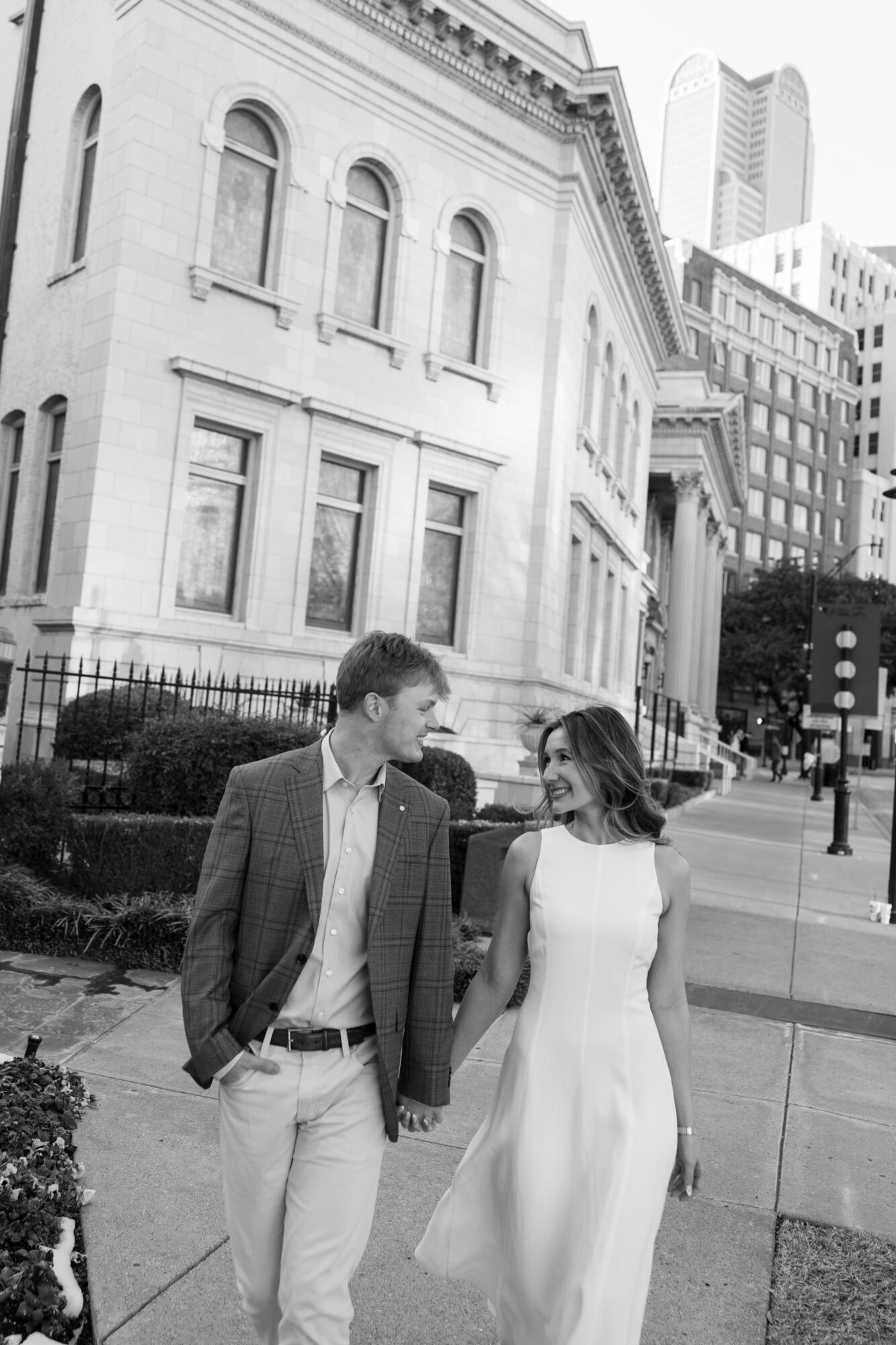 A man and woman holding hands walking on a city sidewalk with historic and modern buildings in the background.