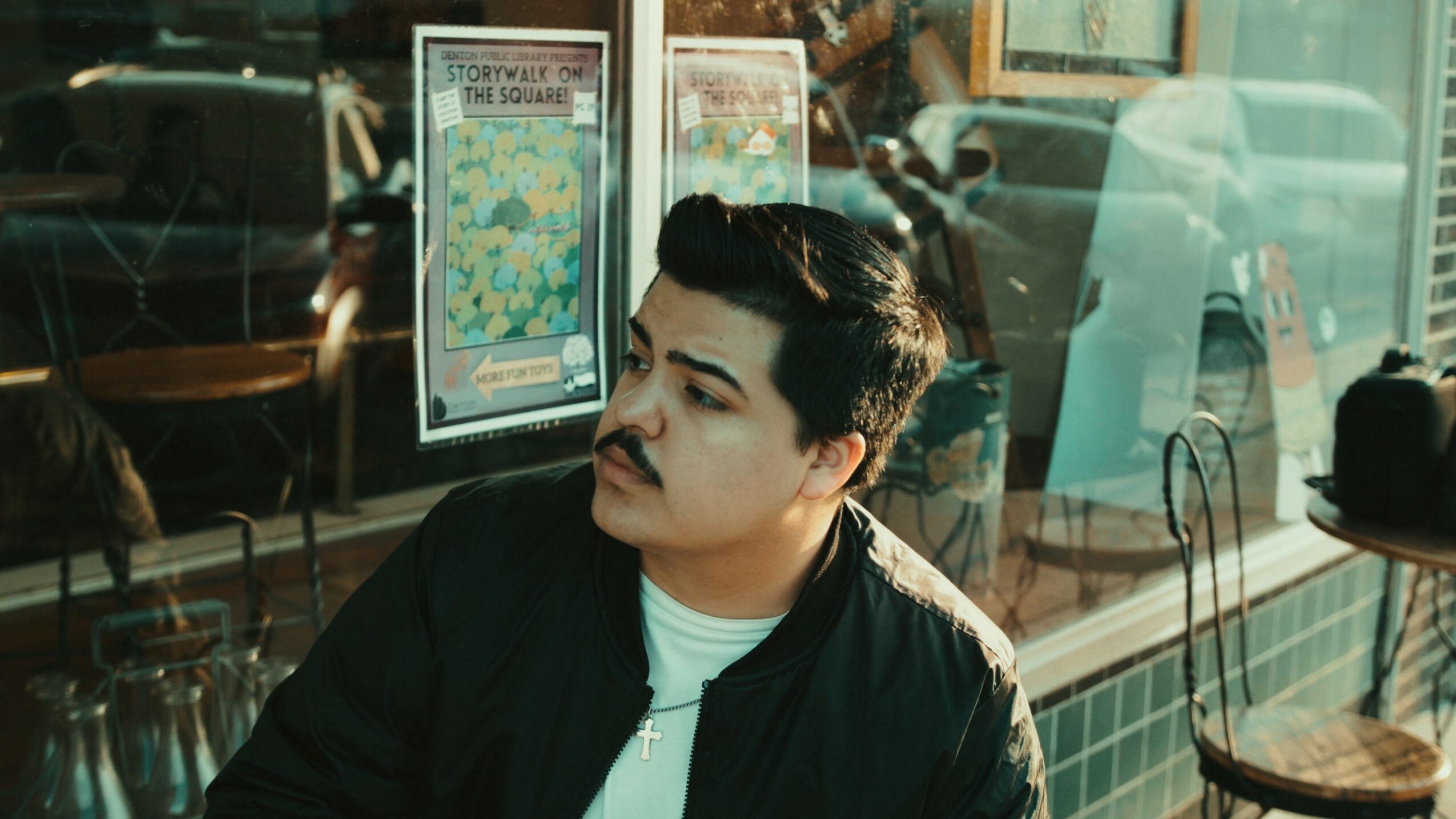 Man with dark hair and mustache sitting indoors, looking to the side, with posters and reflections in the background.