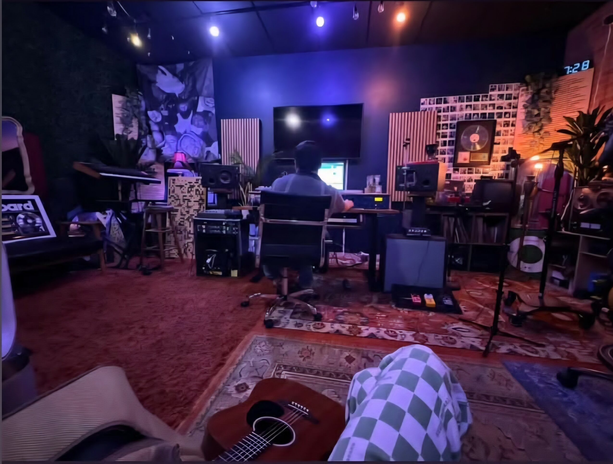 Person working at a music studio with equipment, guitar in foreground, colorful wall decorations, and plants.