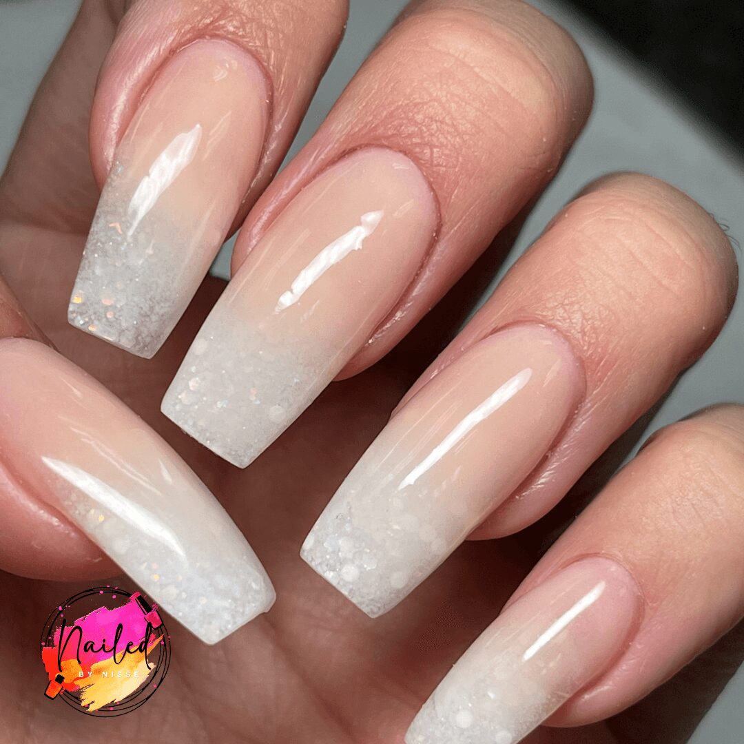 Close-up of five fingers with long, square-shaped nails painted in a gradient white design with glitter at tips.