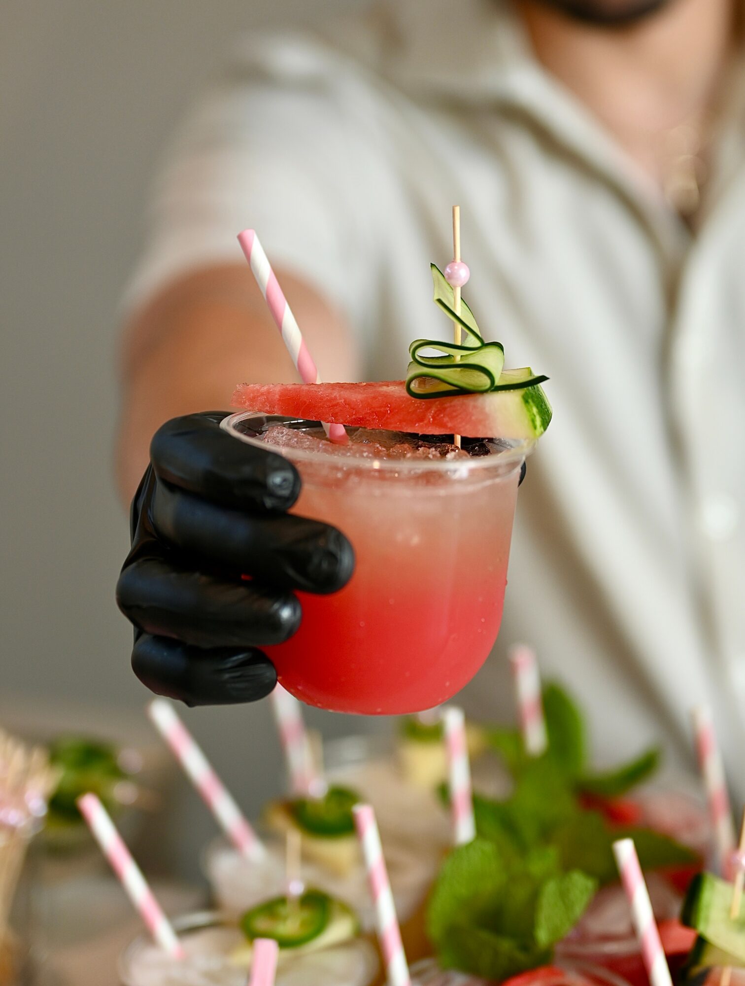 Person wearing black glove holding a pink cocktail with garnishes, with a tray of similar drinks in front.