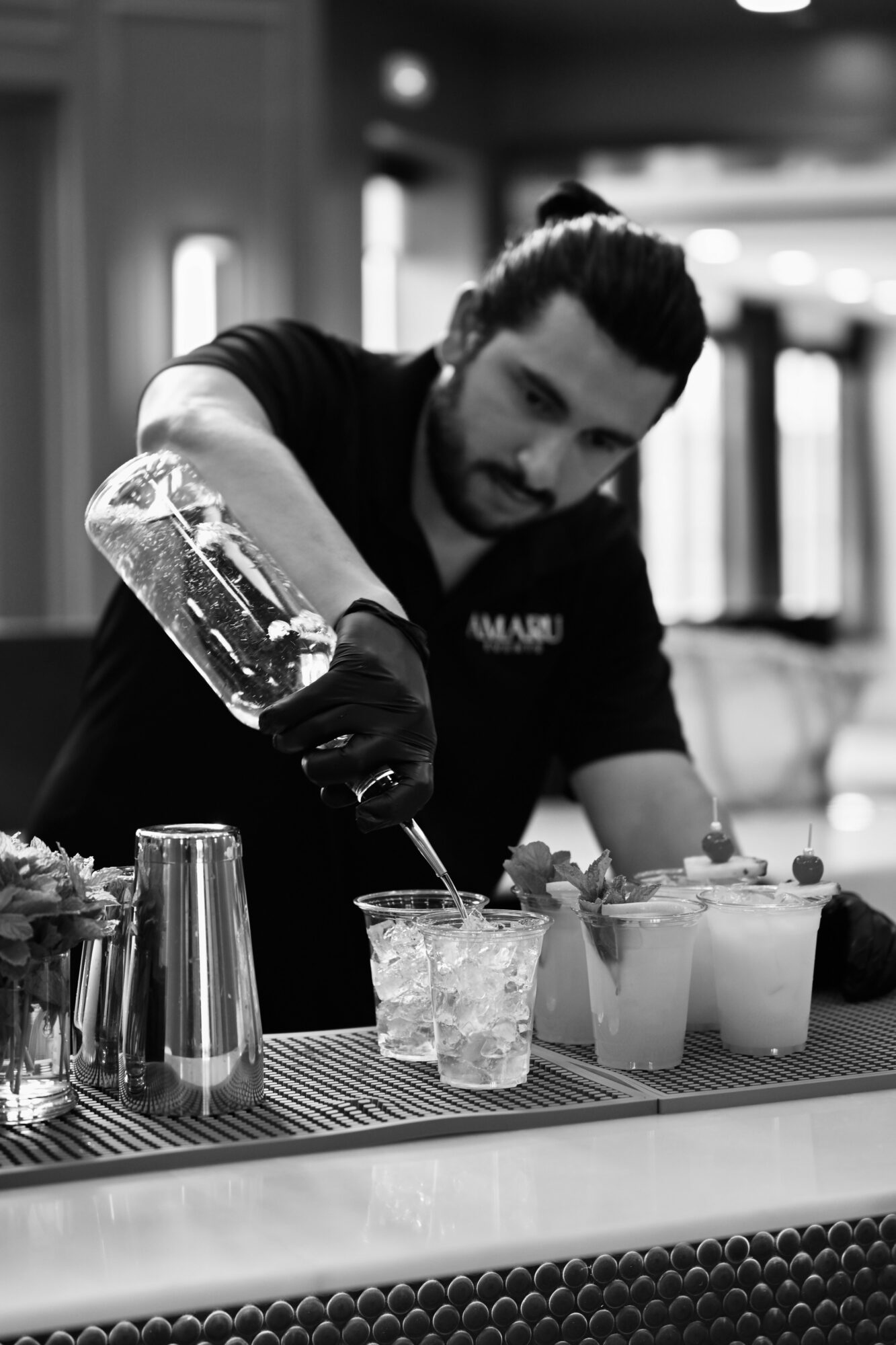 Man pouring a drink into a glass at a bar counter with various bottles and glasses nearby.