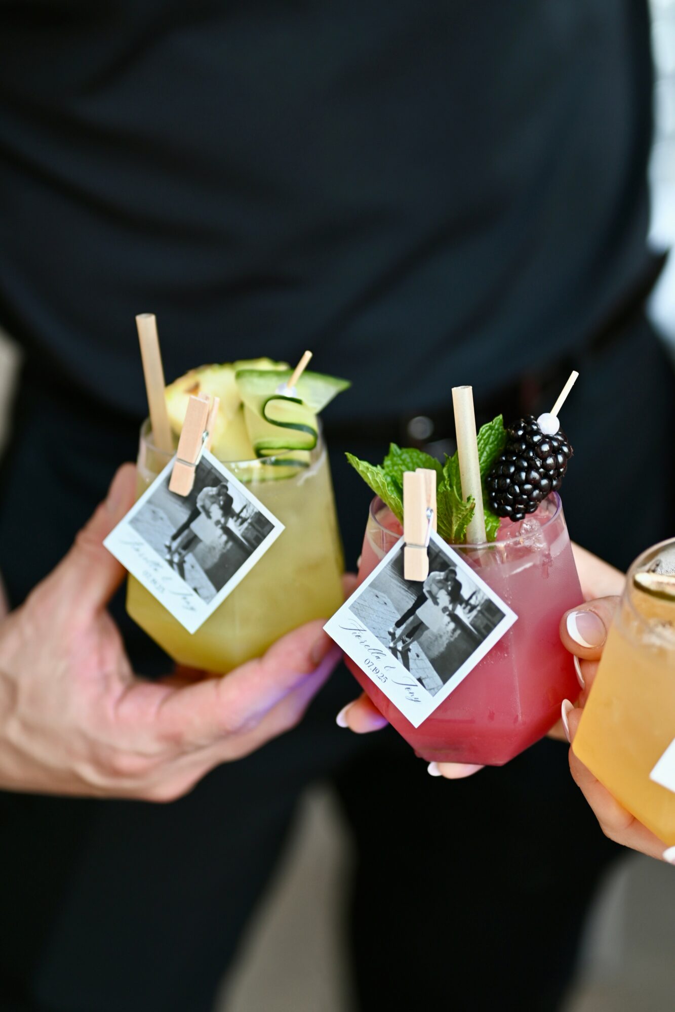 Two colorful drinks with garnishes and small photos attached, held by a person in dark clothing.