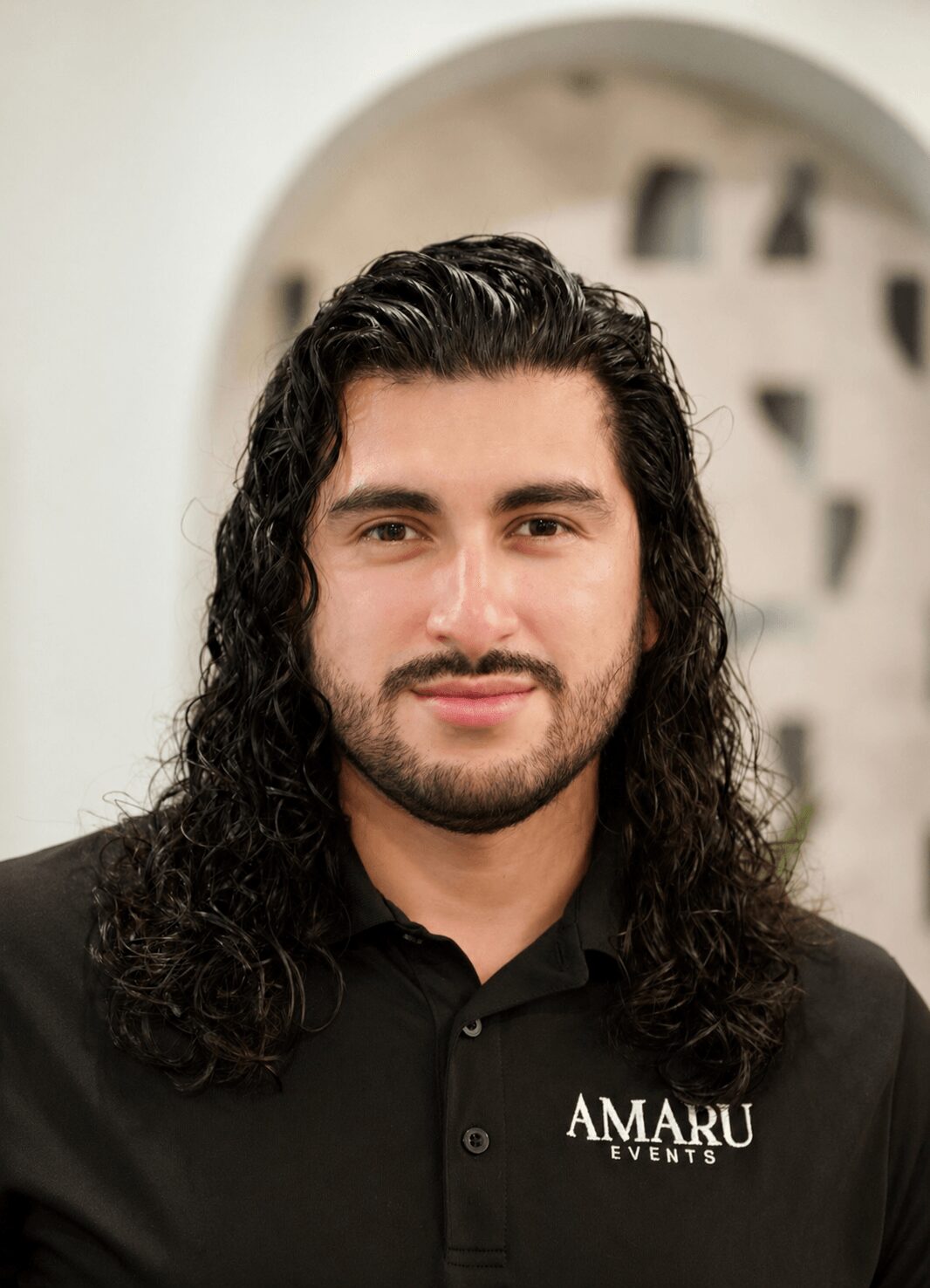 Man with long, curly dark hair and beard wearing a black shirt with 'Amaru Events' logo.