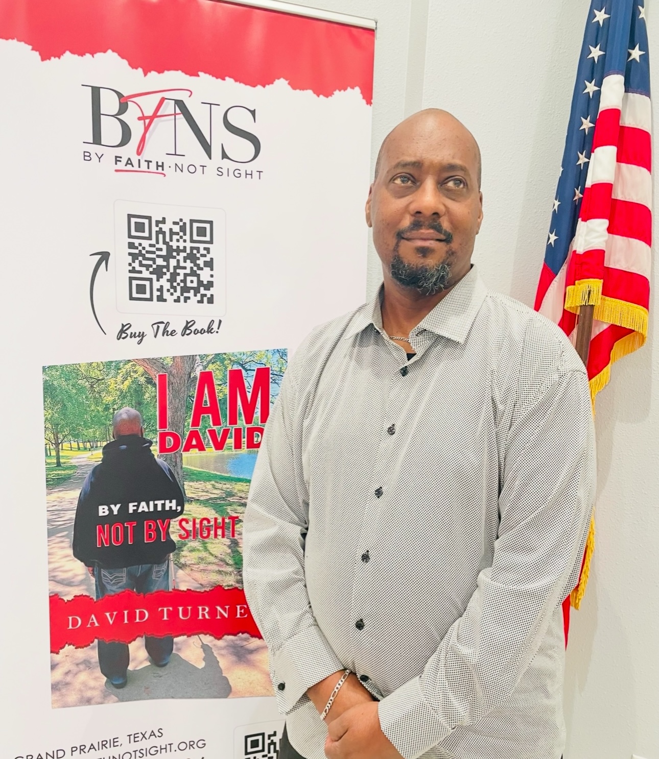 Man in light gray shirt standing indoors near an American flag and a banner with QR code and text.
