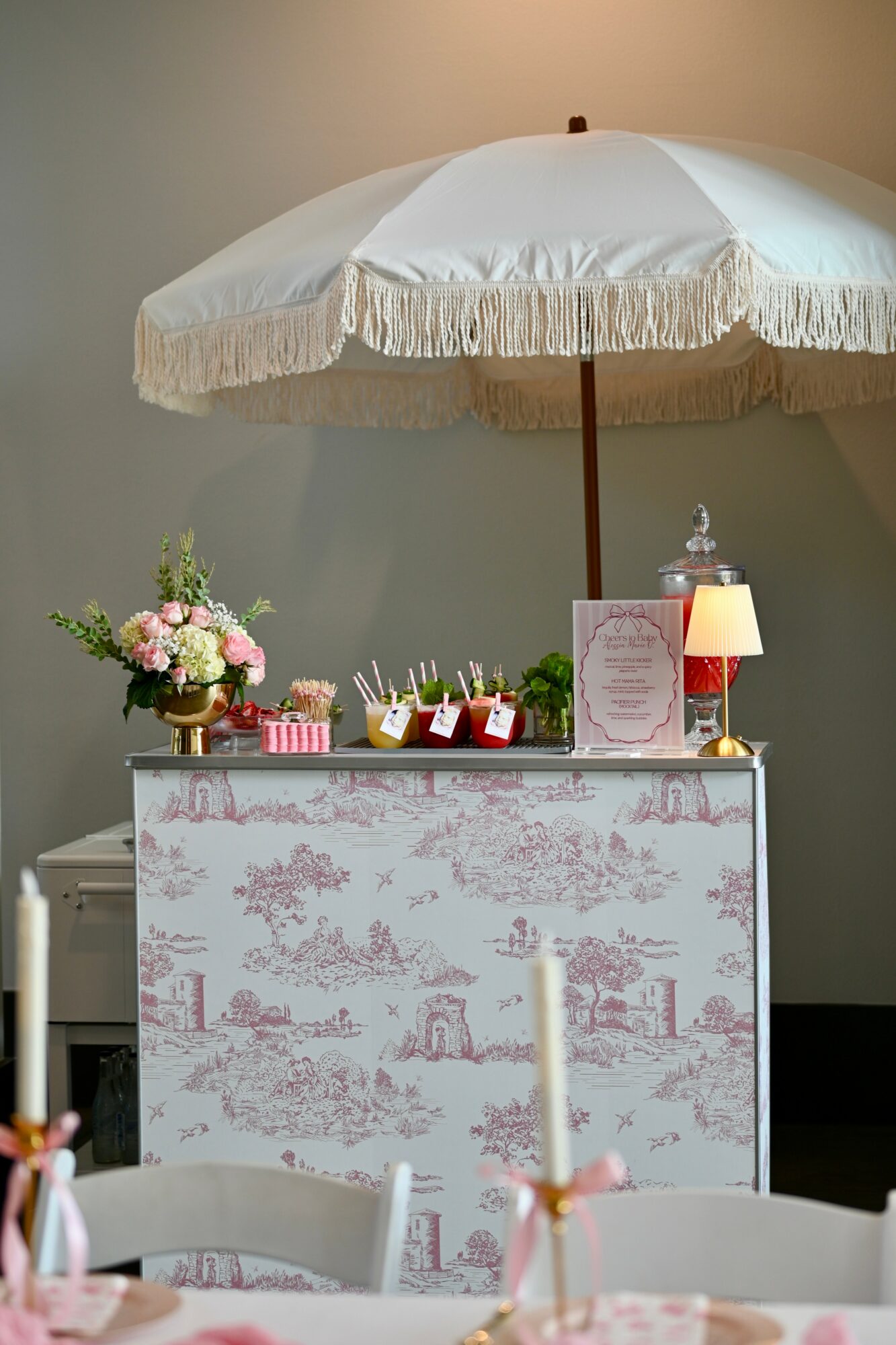 Bar counter with pink and white decor, flowers, small lamps, and umbrellas, set against a plain wall.