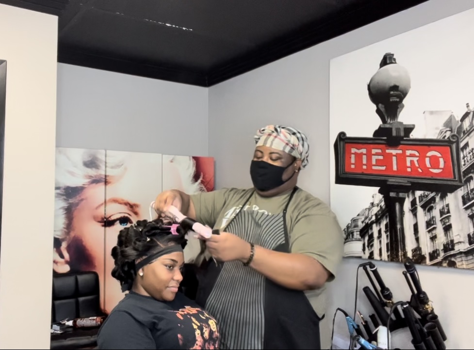 Person getting hair styled by a stylist wearing a mask and head covering in a salon.
