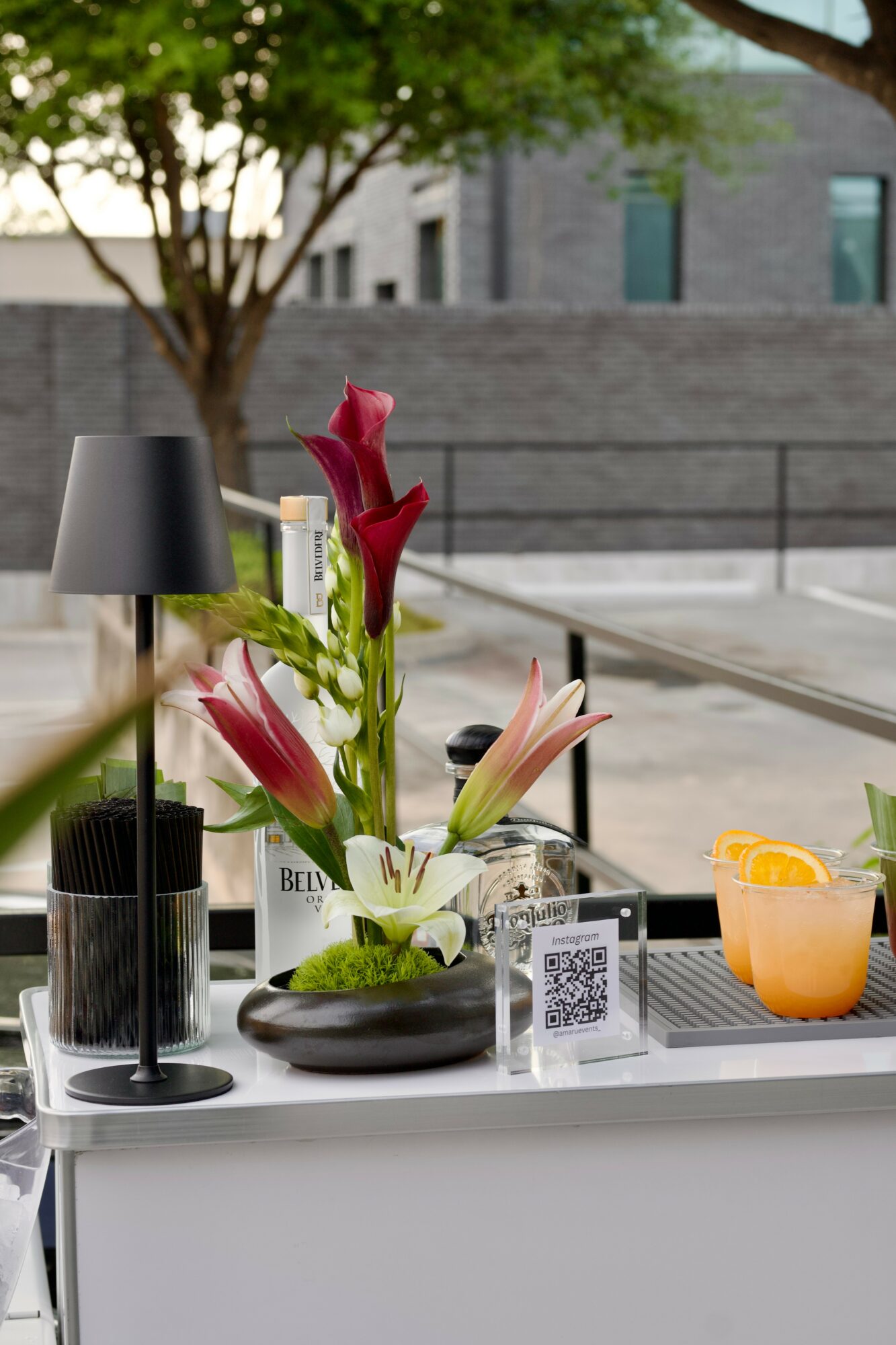 Table with flowers, a black lamp, a QR code, and a drink outdoors with a tree and building in background.