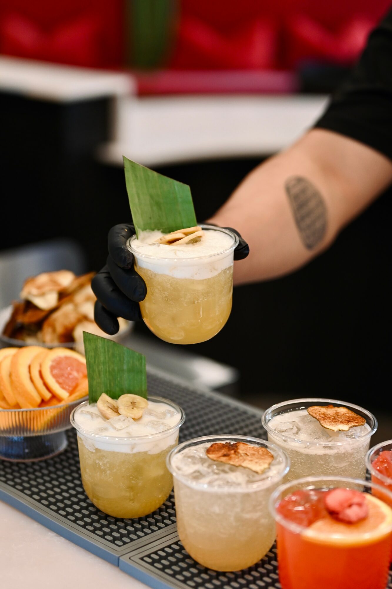 Person holding a cocktail glass with a green garnish, with three similar drinks on a counter, some with fruit toppings.