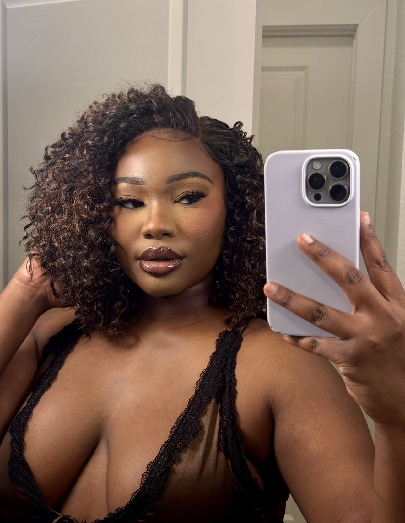 Woman with curly hair taking a mirror selfie, wearing a black top, in a room with white walls and door.