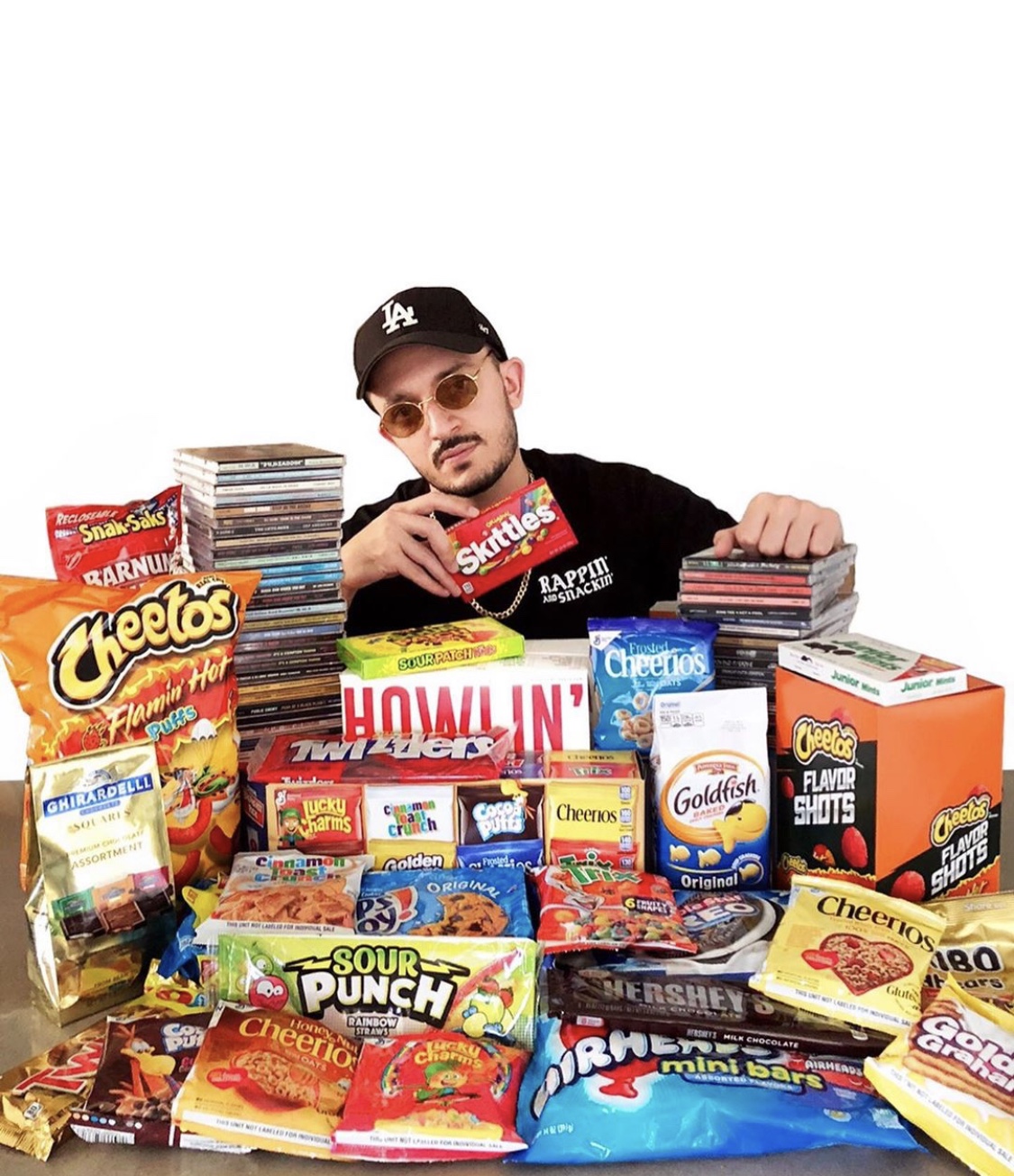Young man wearing sunglasses and a cap, surrounded by snacks and candy on a table.