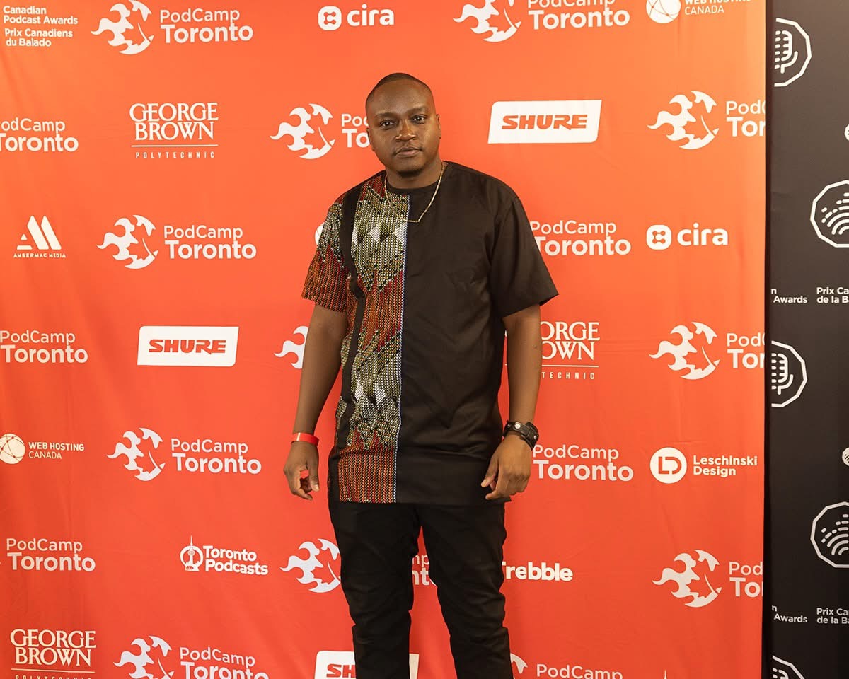 Man standing in front of a red backdrop with logos and text, wearing a black and patterned shirt and black pants.