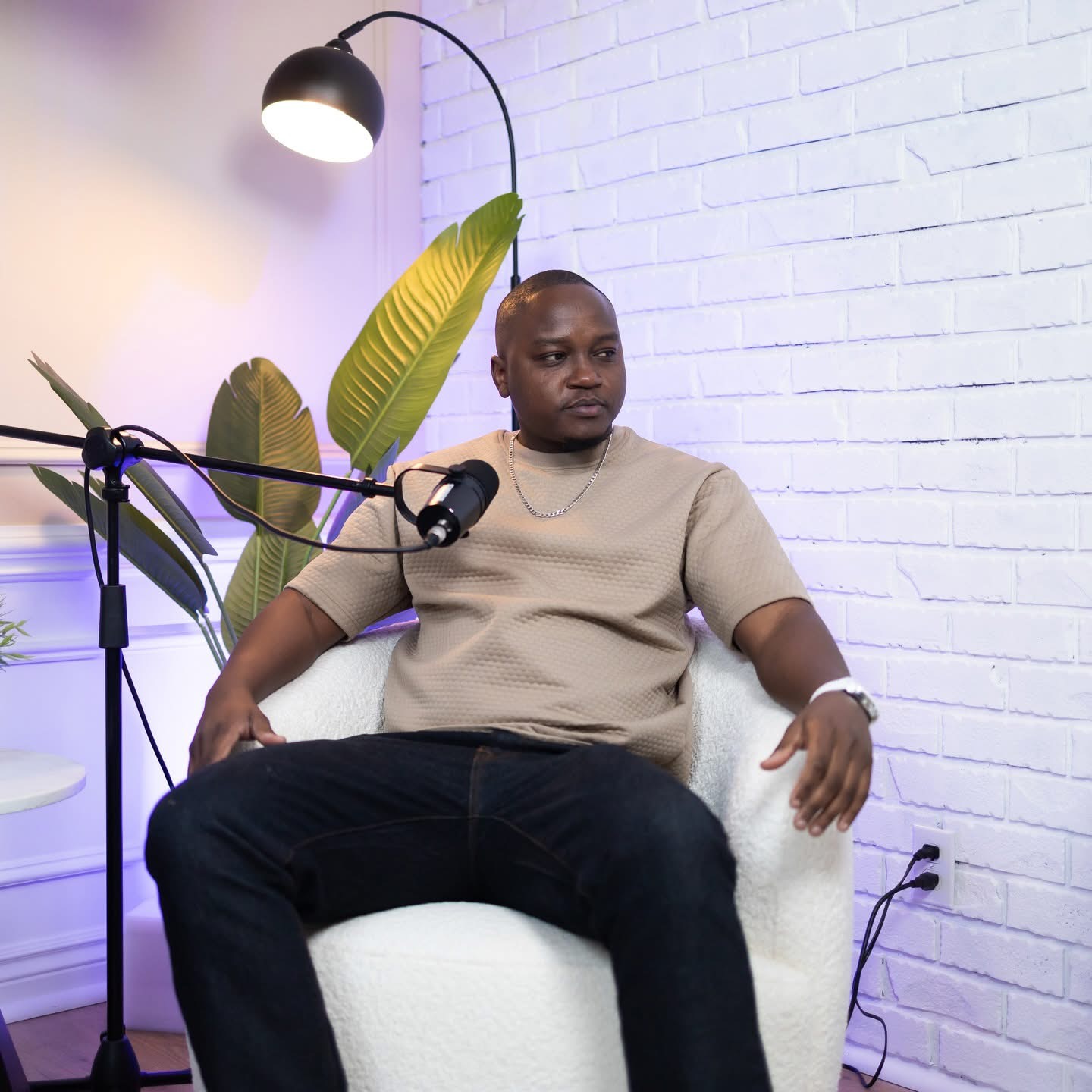 Man sitting in a white chair with a microphone, plants, and a lamp in a room with white brick wall.