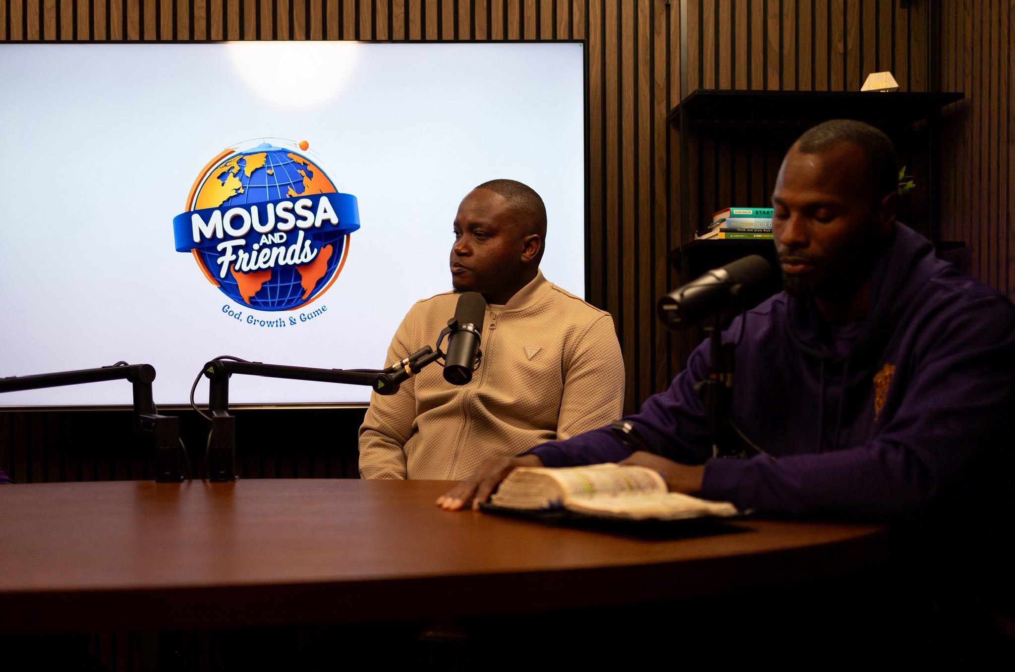 Two men sit at a table with microphones, one reading a book, in a room with wood-paneled walls and a screen displaying a logo.