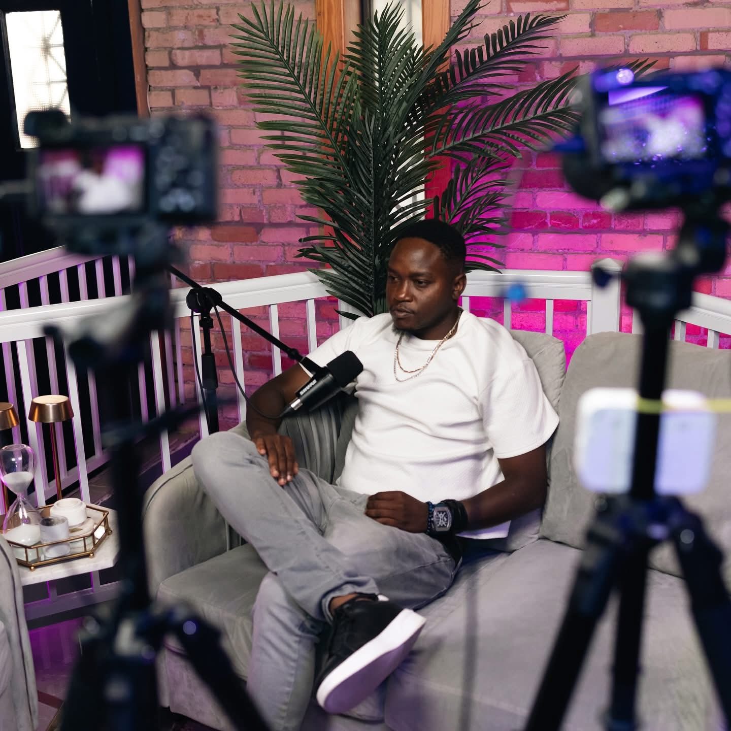 Man sitting on a sofa with cameras recording him, brick wall and plant in background.