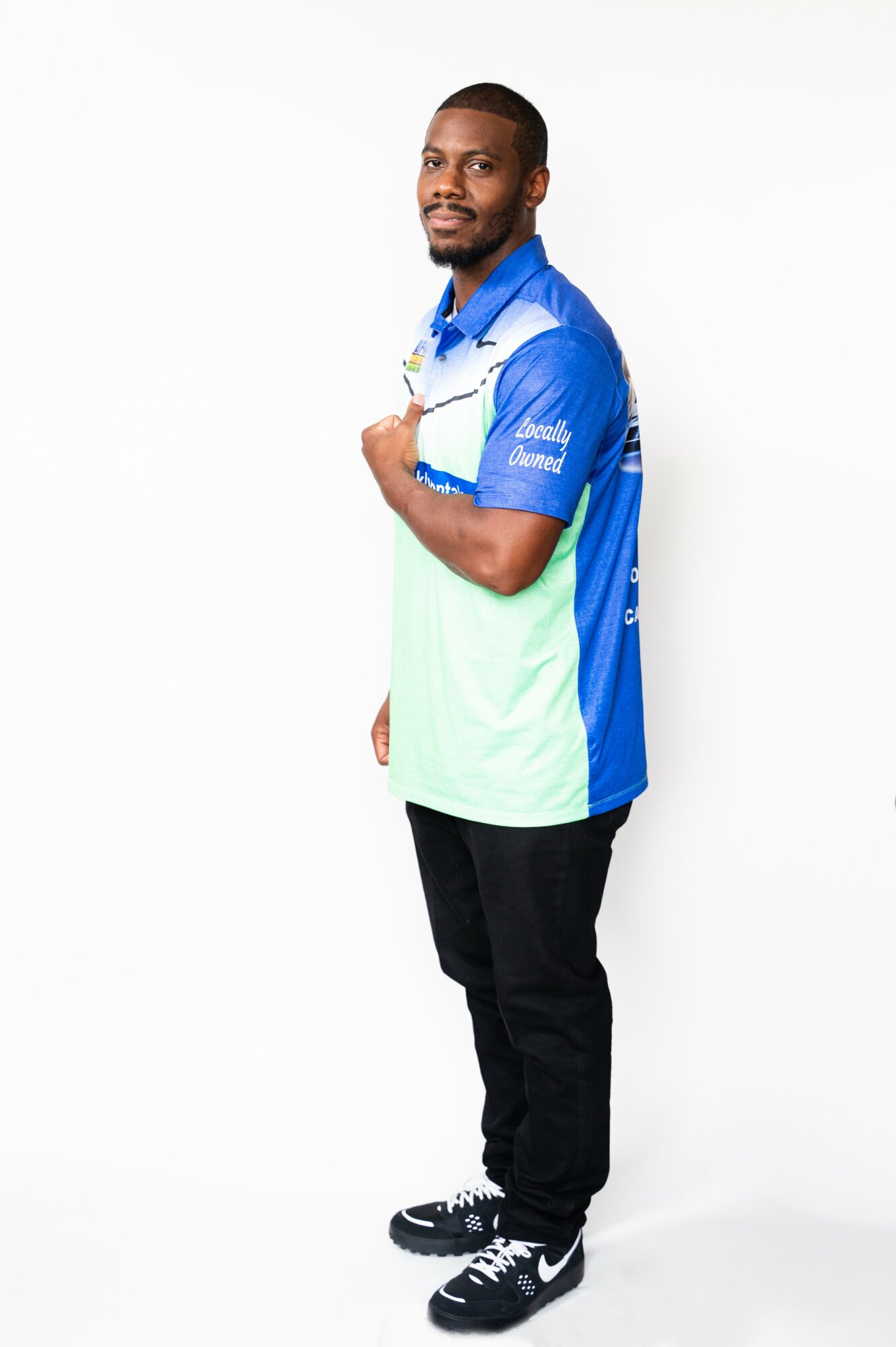 Man standing with arms crossed, wearing a blue and white sports shirt and black pants, against a white background.