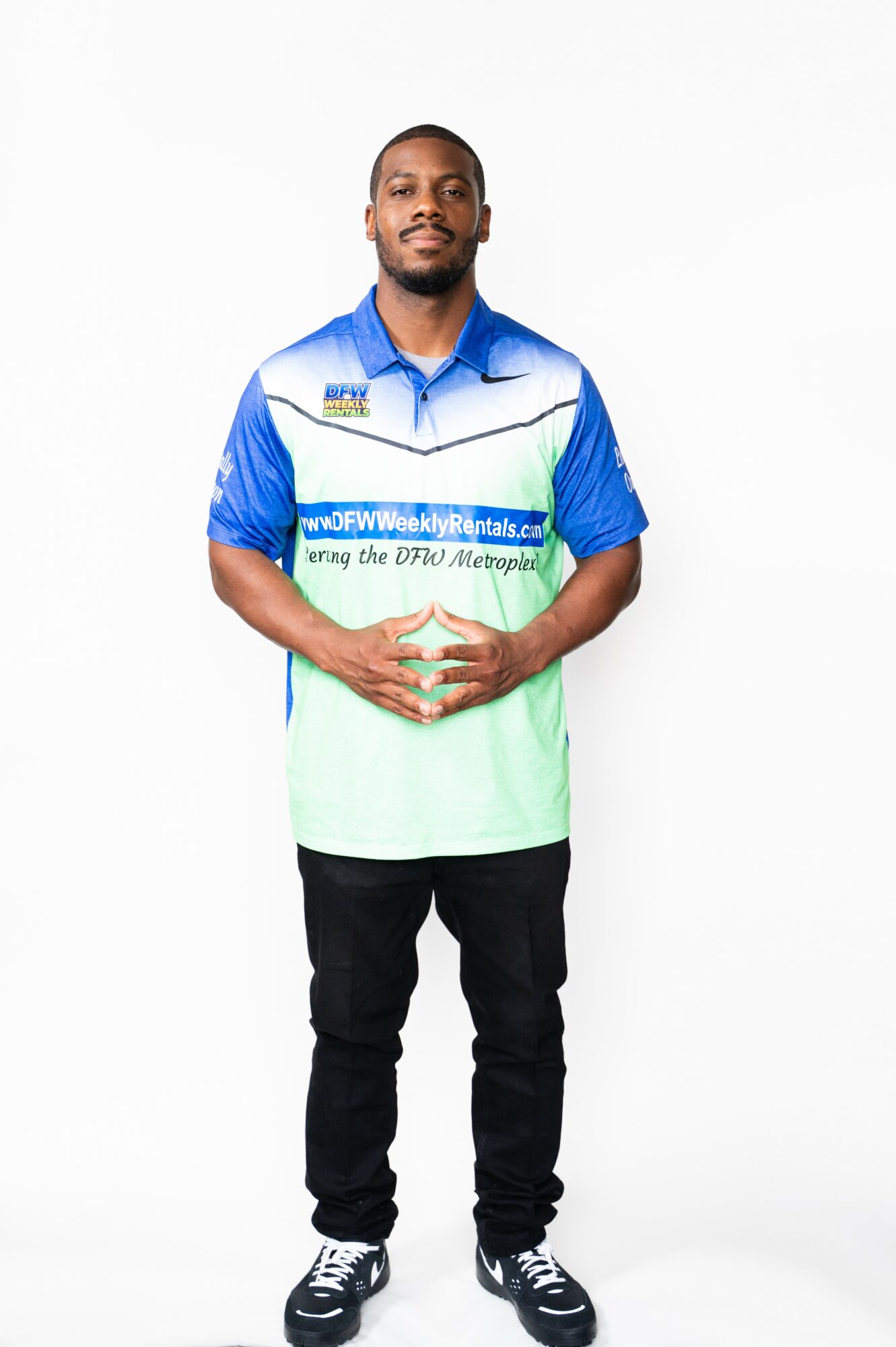Young man standing with hands clasped, wearing a blue and green sports shirt, black pants, and sneakers.