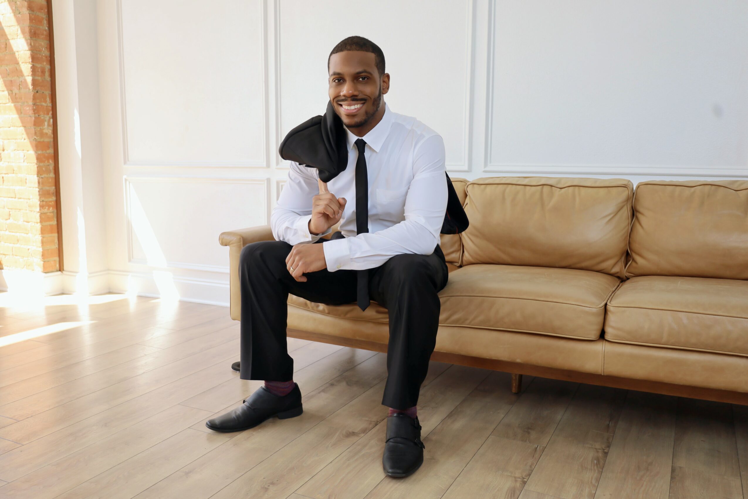 Man sitting on a beige sofa, smiling, holding a jacket over his shoulder, in a bright room with wooden floor.