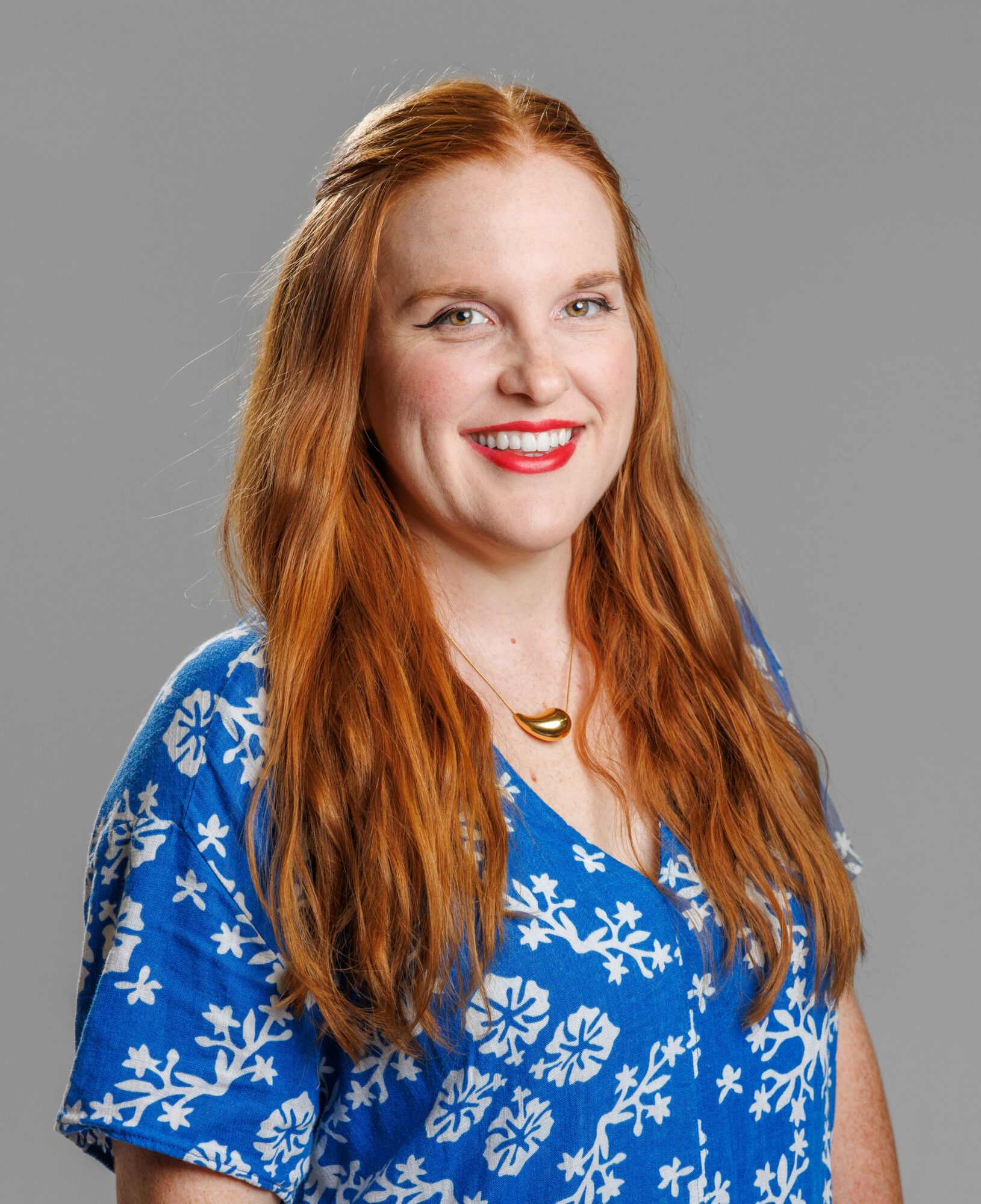 Smiling woman with long red hair wearing a blue floral shirt and a necklace, against a gray background.
