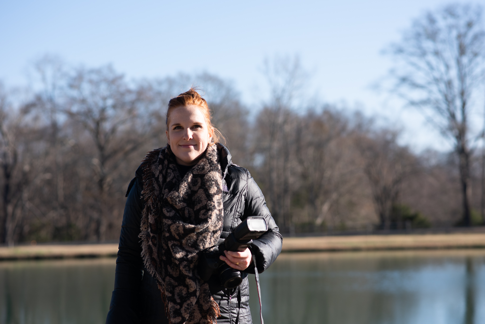 Woman standing outdoors near a lake with trees in the background, holding a camera, wearing a scarf and jacket.