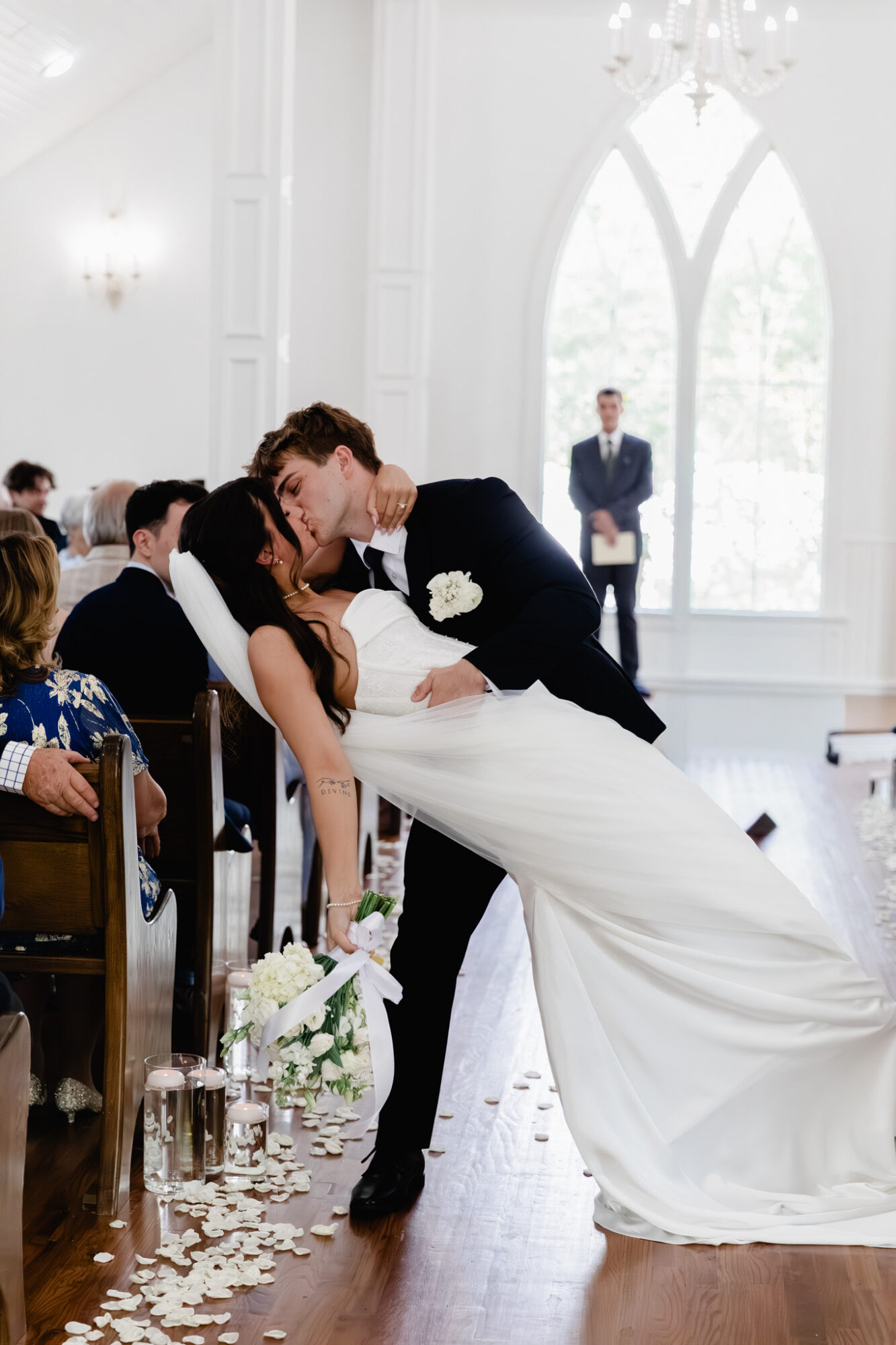 Groom dips bride for a kiss during wedding ceremony in bright, elegant hall with large arched window.