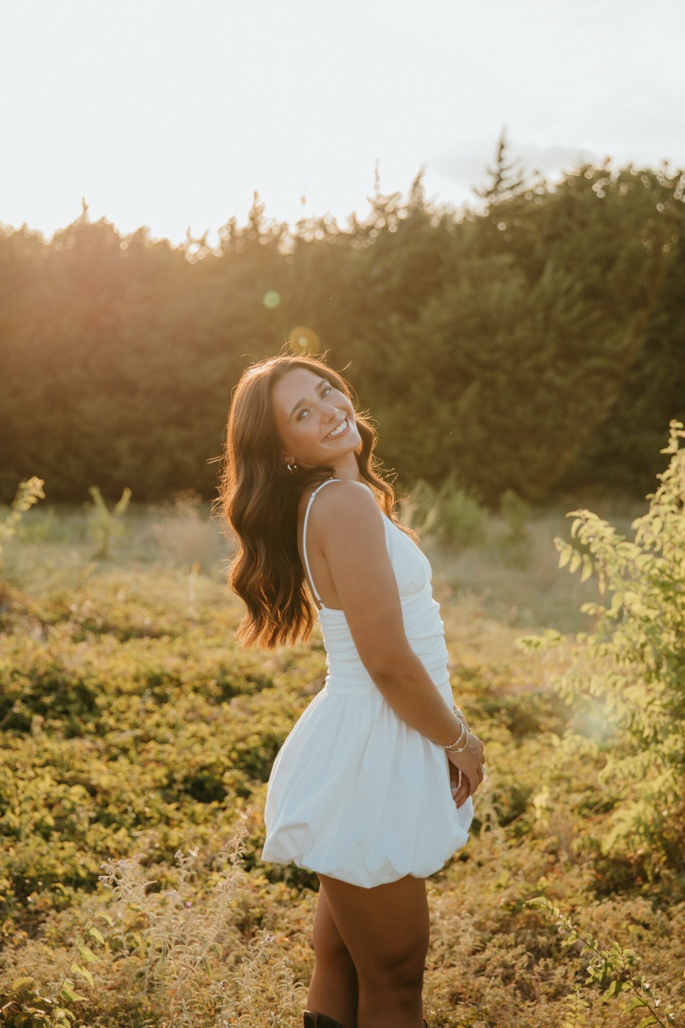 Young woman in white dress standing outdoors in sunlight, with trees in background.