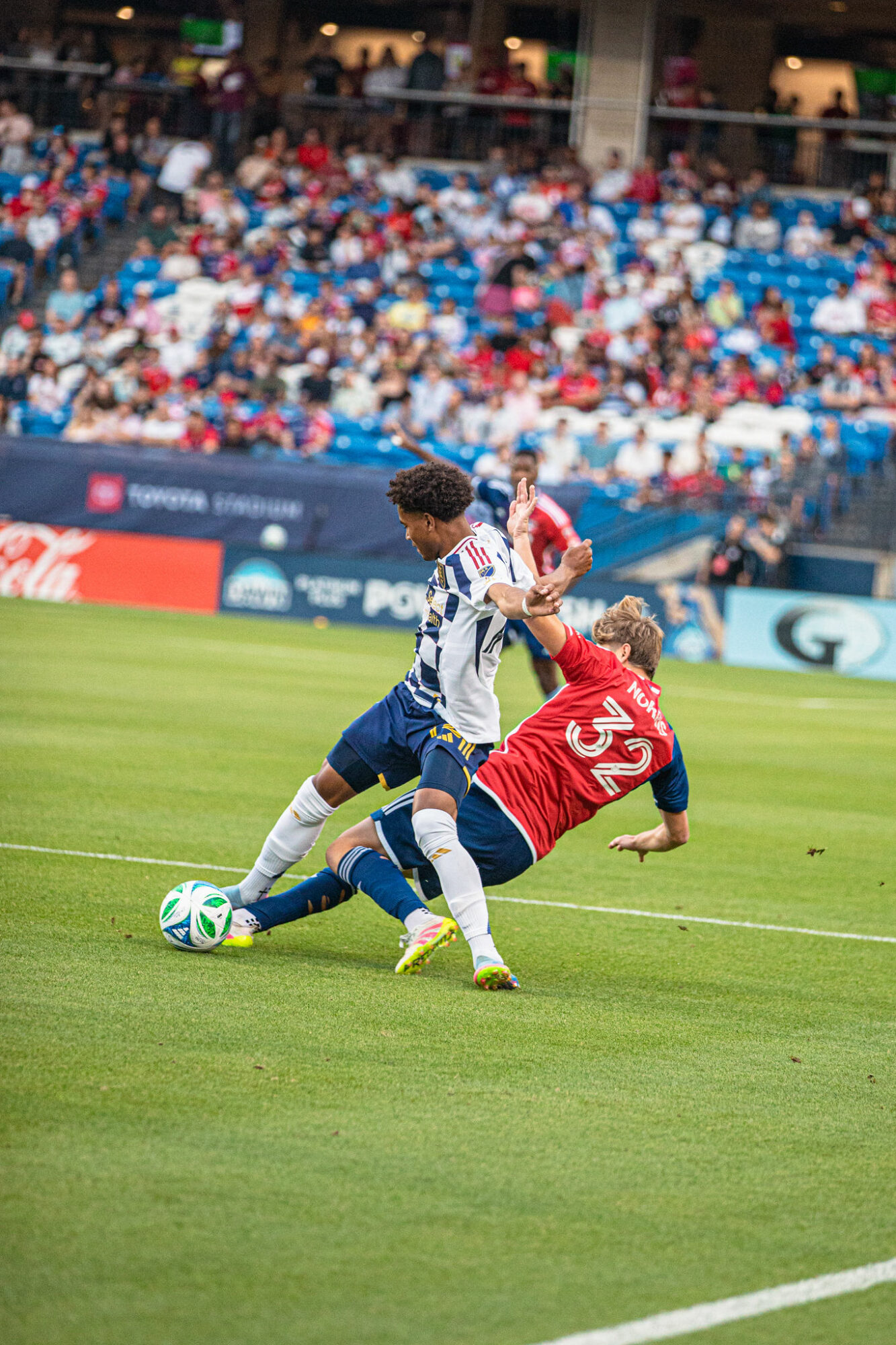 Two soccer players compete for the ball on a green field with a crowd in the background.