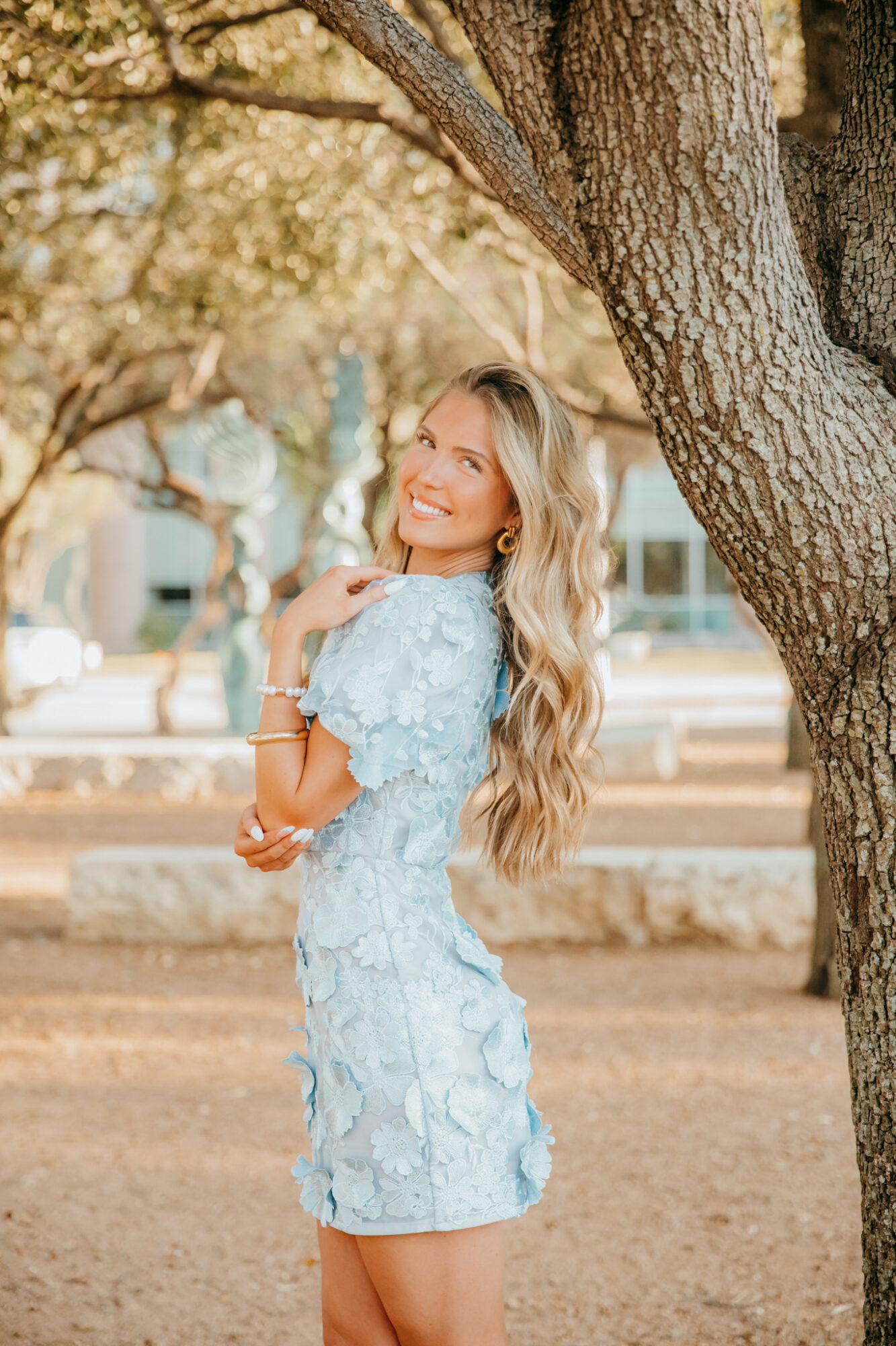 Young woman with long wavy hair smiling outdoors near a tree, wearing a light blue dress.