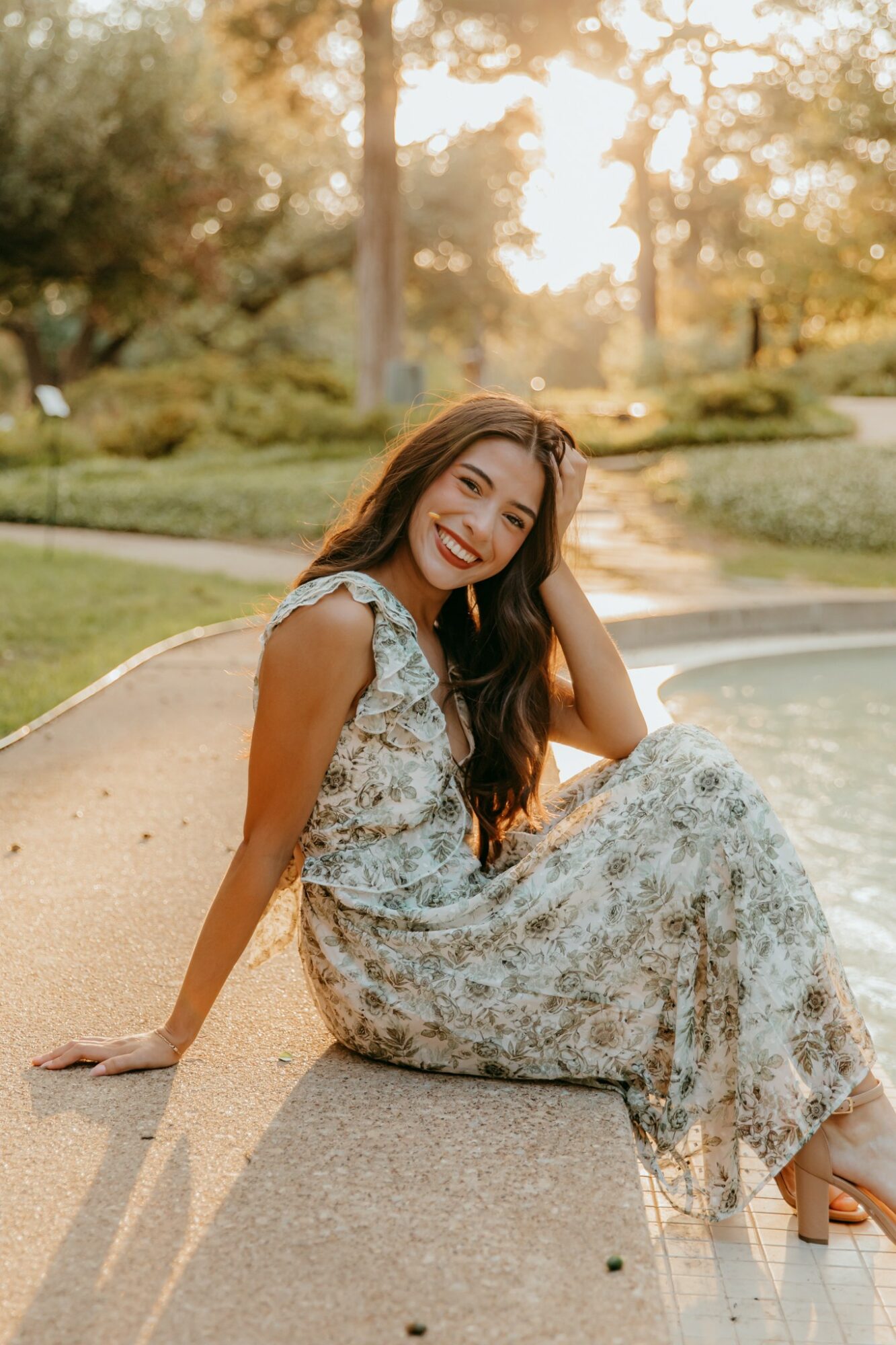 Woman sitting by a pool in a floral dress, smiling, with trees and sunlight in the background.
