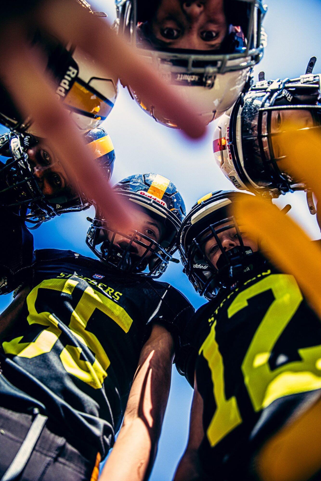 Group of football players in helmets and jerseys huddled together, looking downwards, with a focus on their upper bodies and helmets.