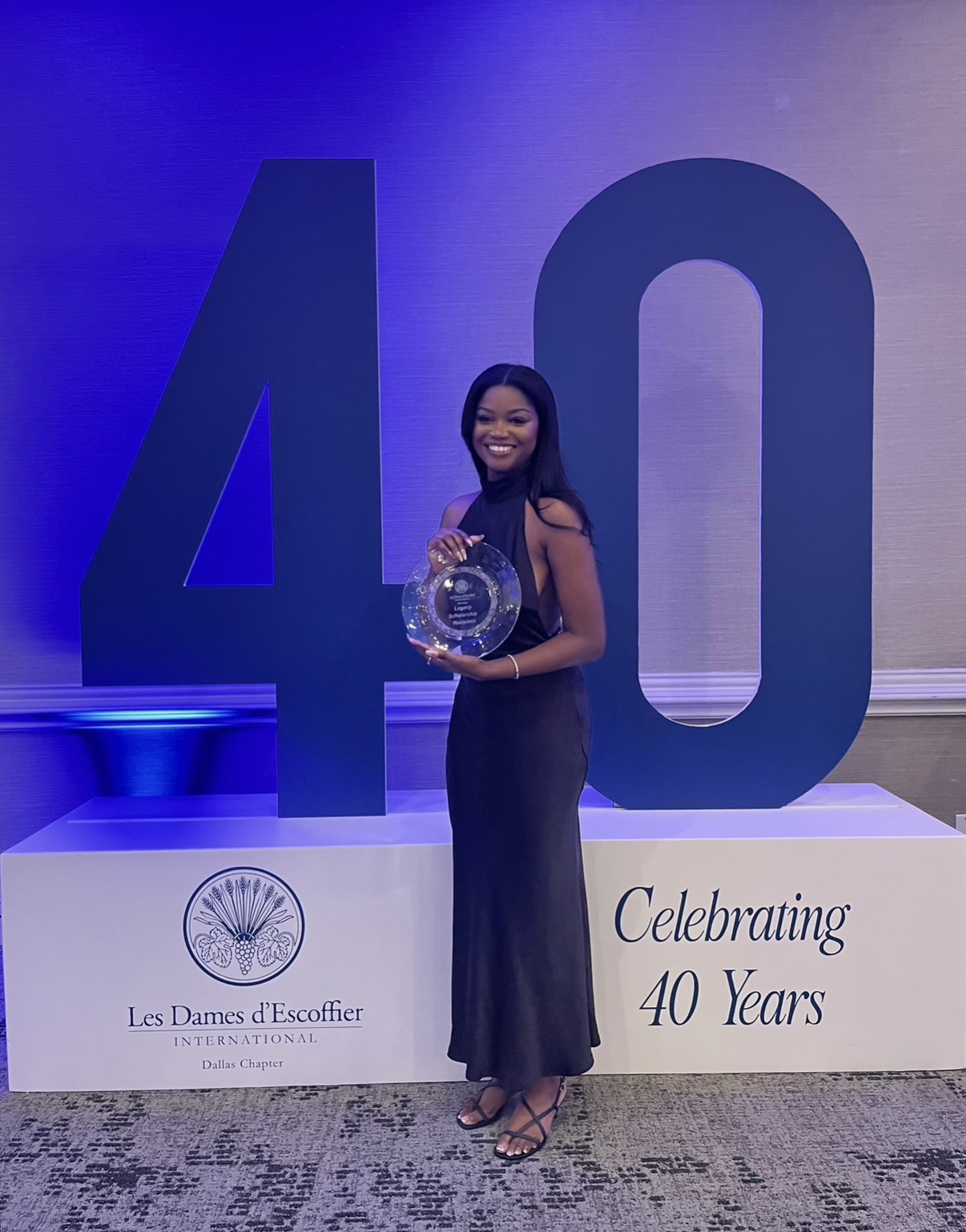 Woman in a long dress holding a glass award, standing in front of large number 40 and celebration sign.
