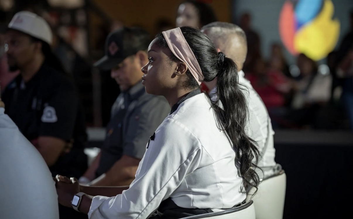 Group of people seated indoors, some wearing uniforms, with a woman in the foreground wearing a headband and white shirt.