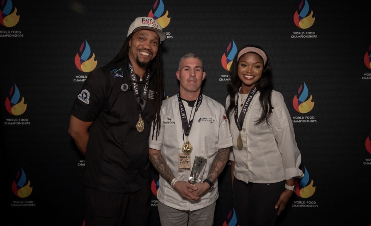 Three people standing together, smiling, with a black background and colorful flame logo pattern.