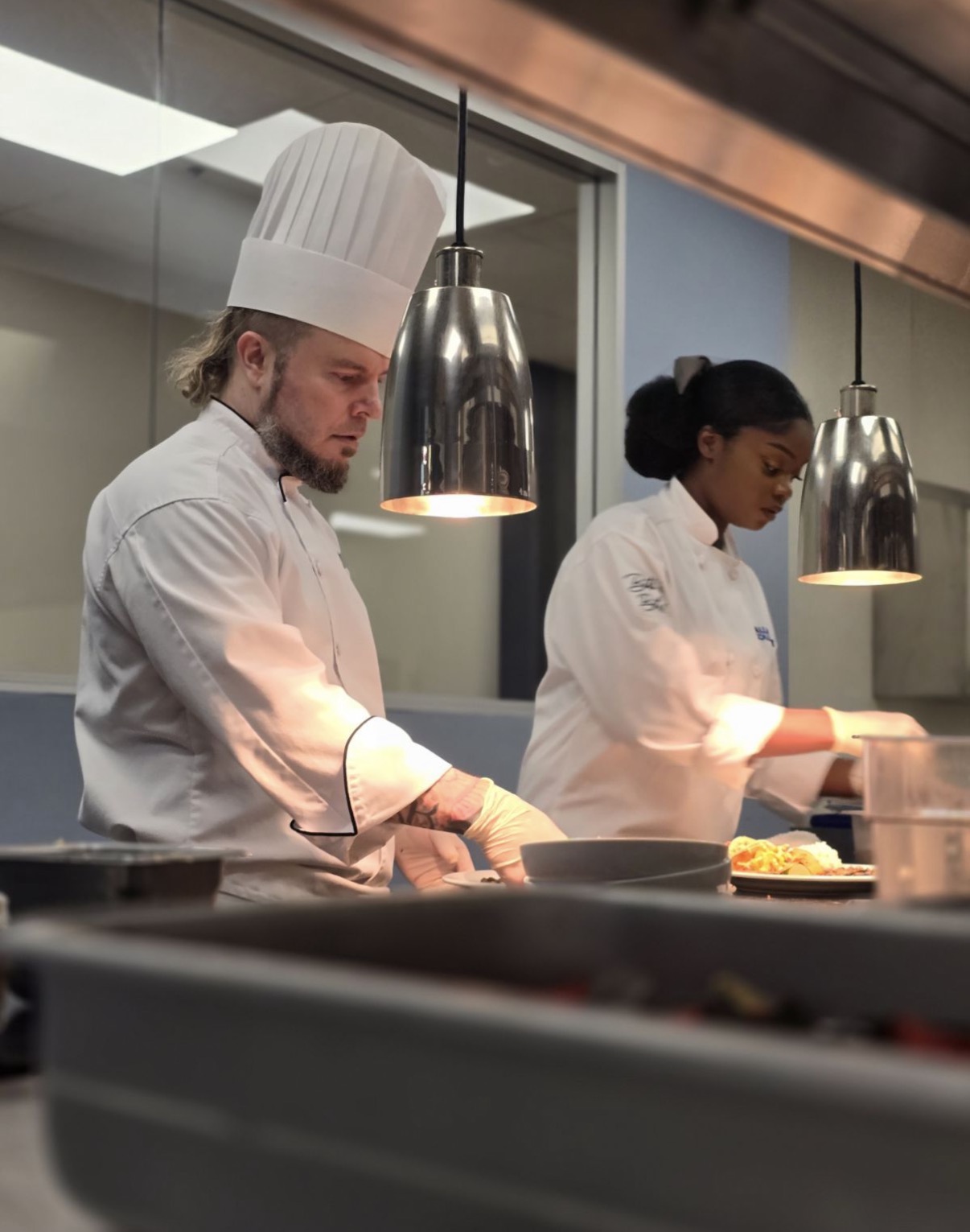 Two chefs in white uniforms and hats work in a professional kitchen with hanging lights.