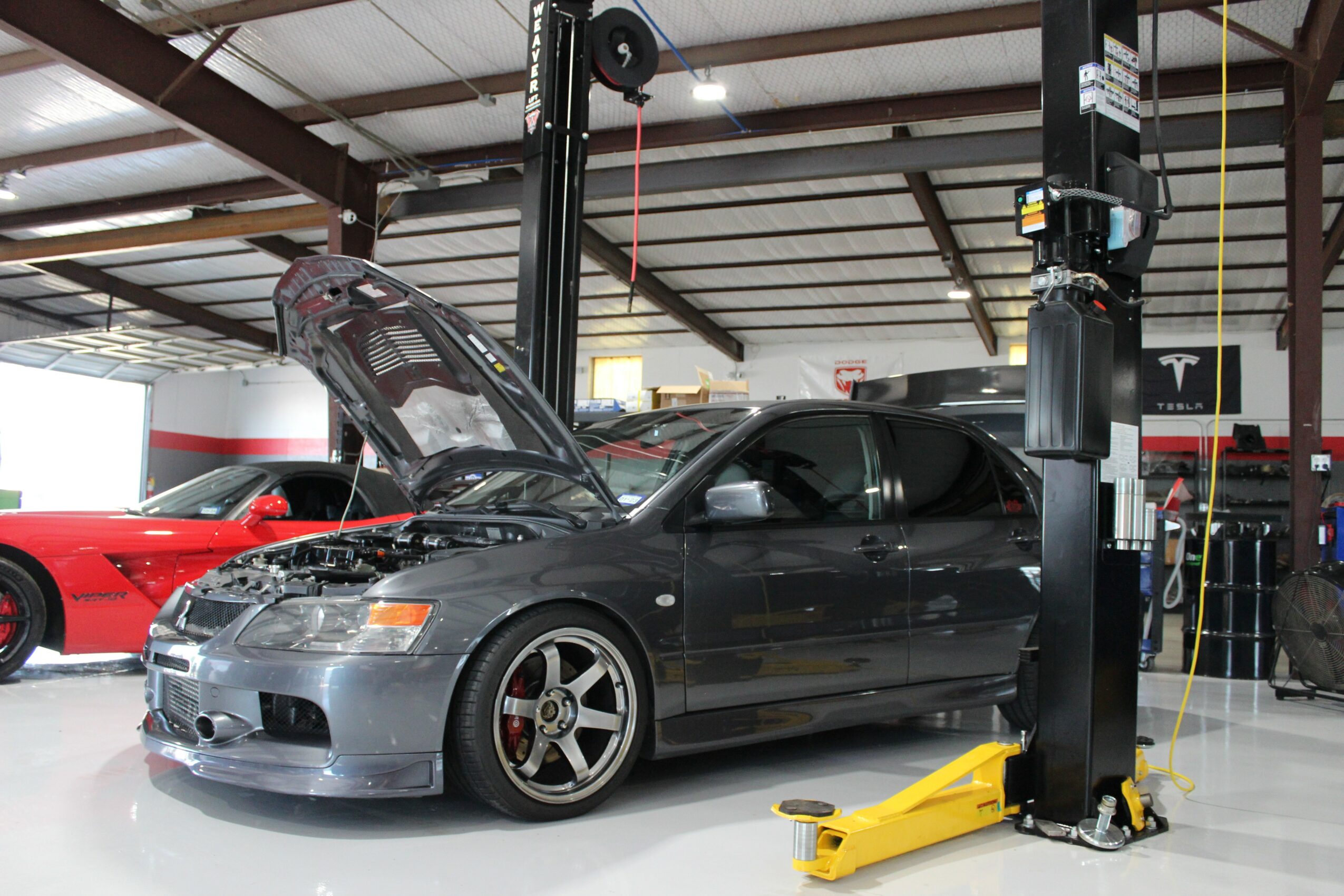 Black car with open hood inside a garage, surrounded by other vehicles and equipment.