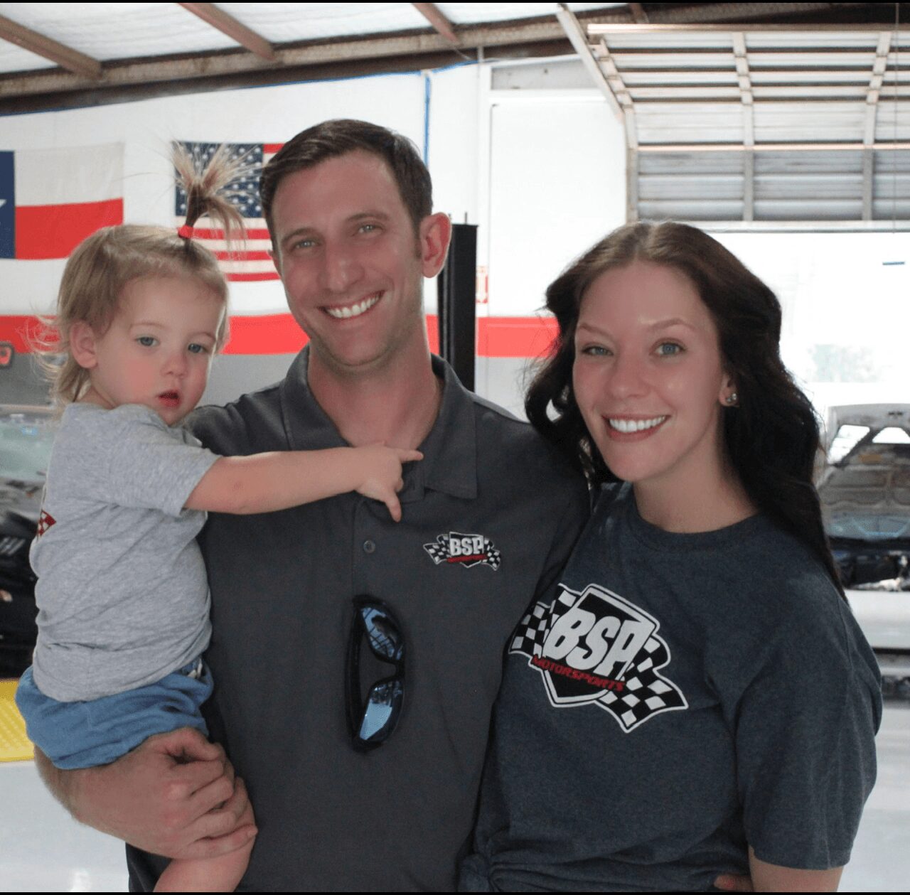 Three people, a man, a woman, and a young girl, smiling indoors with flags in the background.