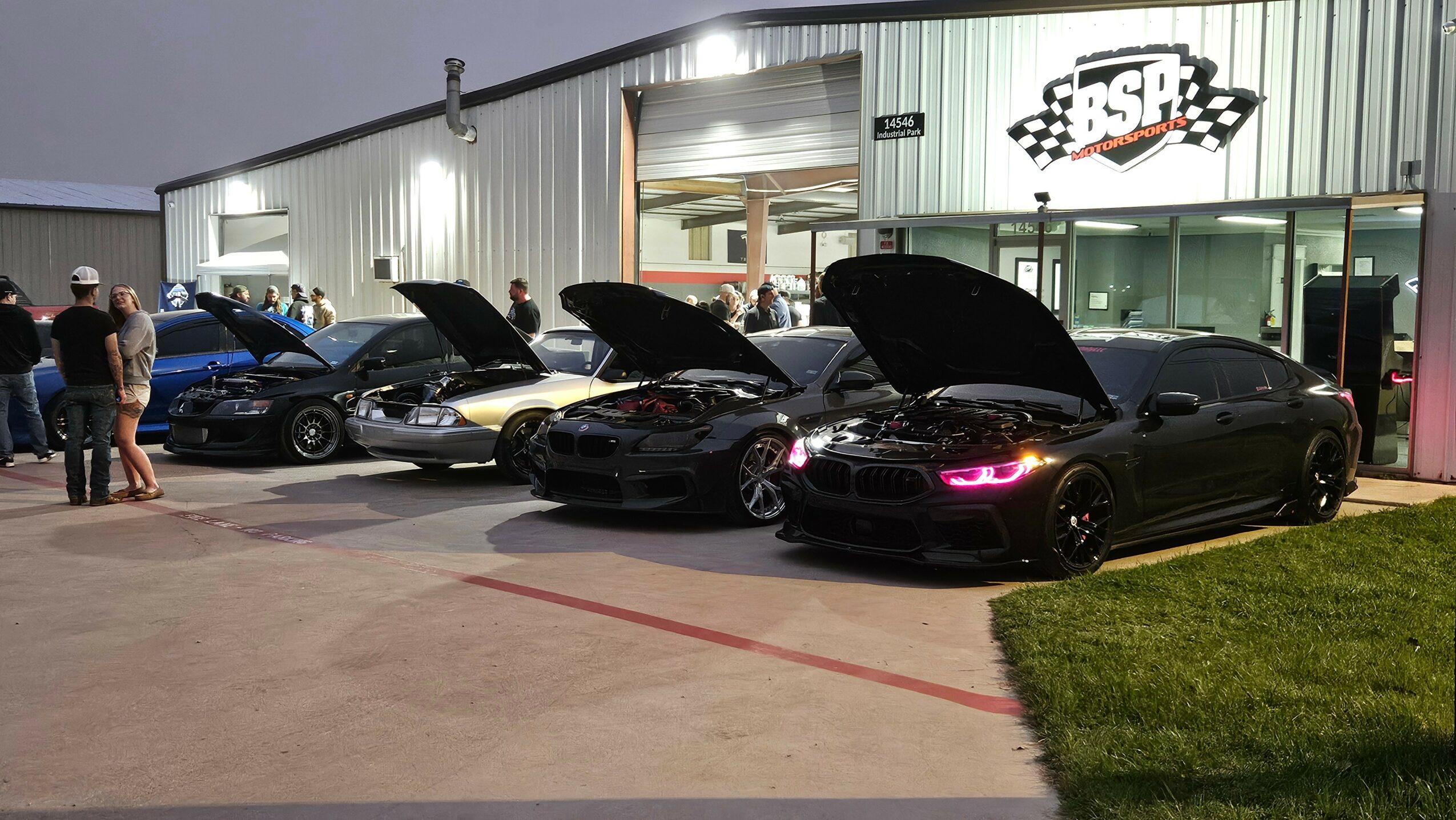 Multiple cars with open hoods parked outside a building with a sign, people standing nearby at night.