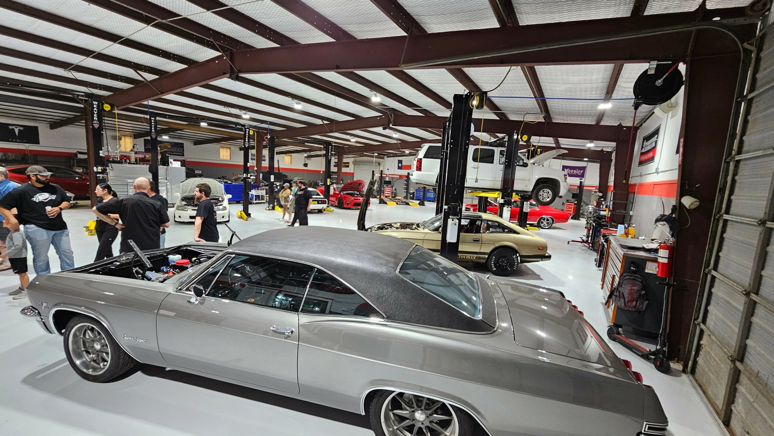 Classic silver car inside a busy garage or showroom with people and vehicles in background.